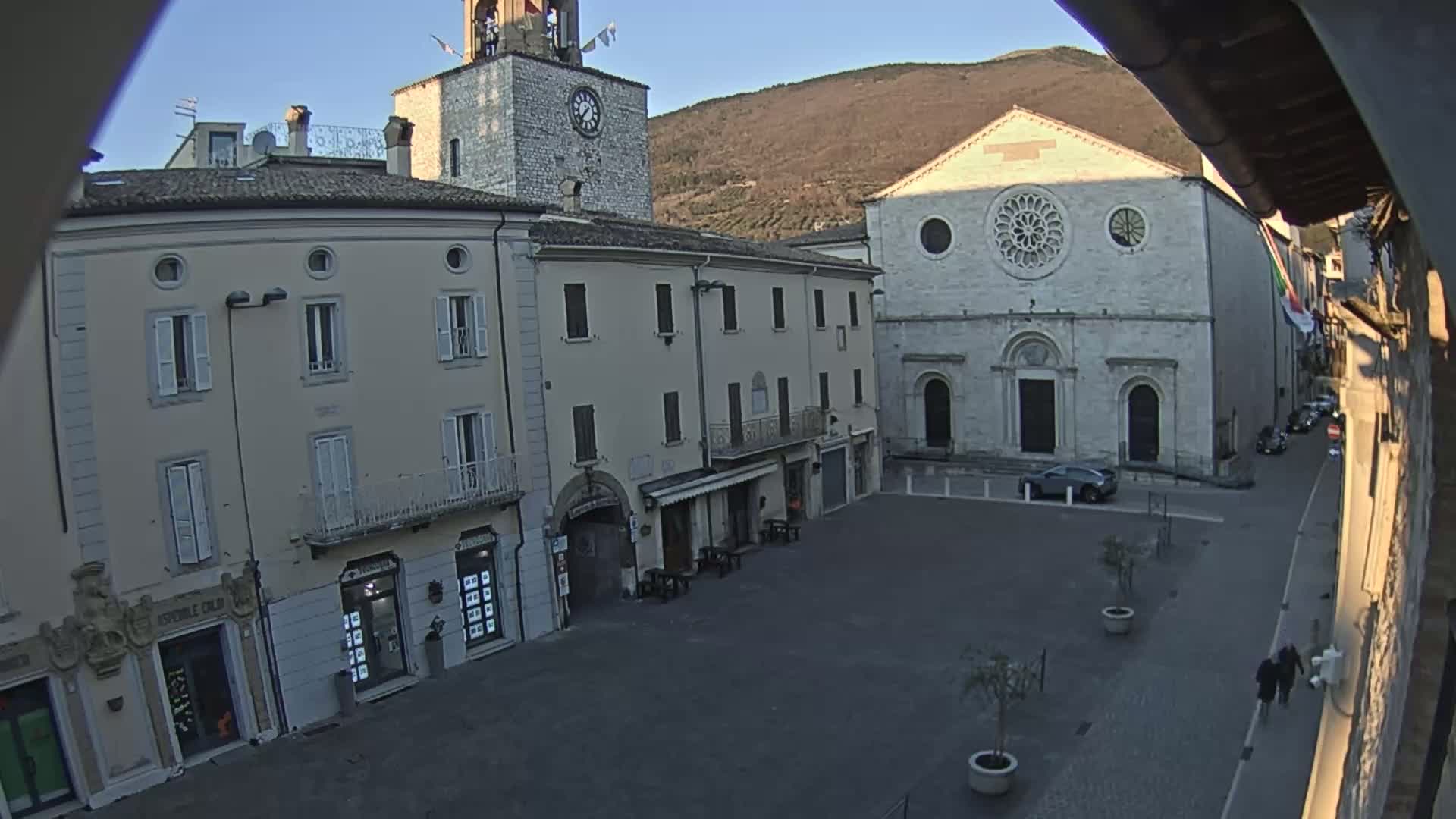 A town square with several buildings, including a church with a rose window and a clock tower, is seen on a clear, sunny day with a mountain in the background.