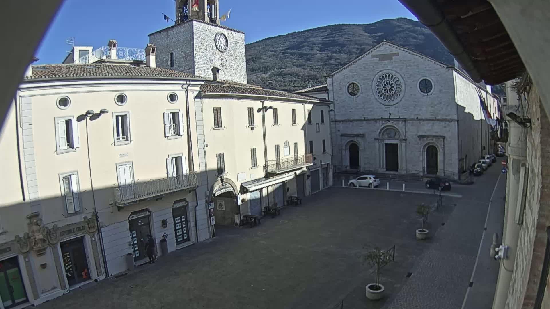 A town square with light-colored buildings, including a church with a rose window and a clock tower, under a clear, sunny sky with mountains in the background.