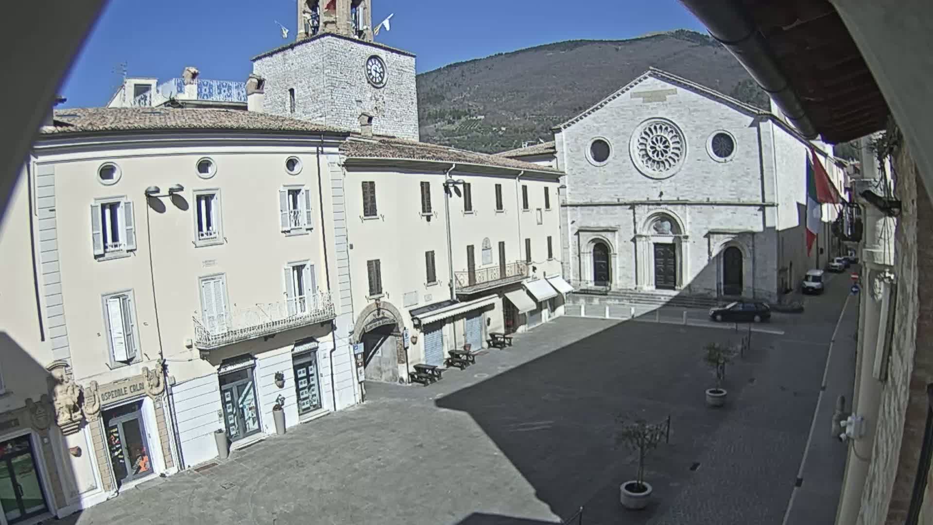 A sunny day reveals a town square with light-colored buildings, including a church with a large rose window, a clock tower, and shops, under a clear blue sky and mountains in the background.