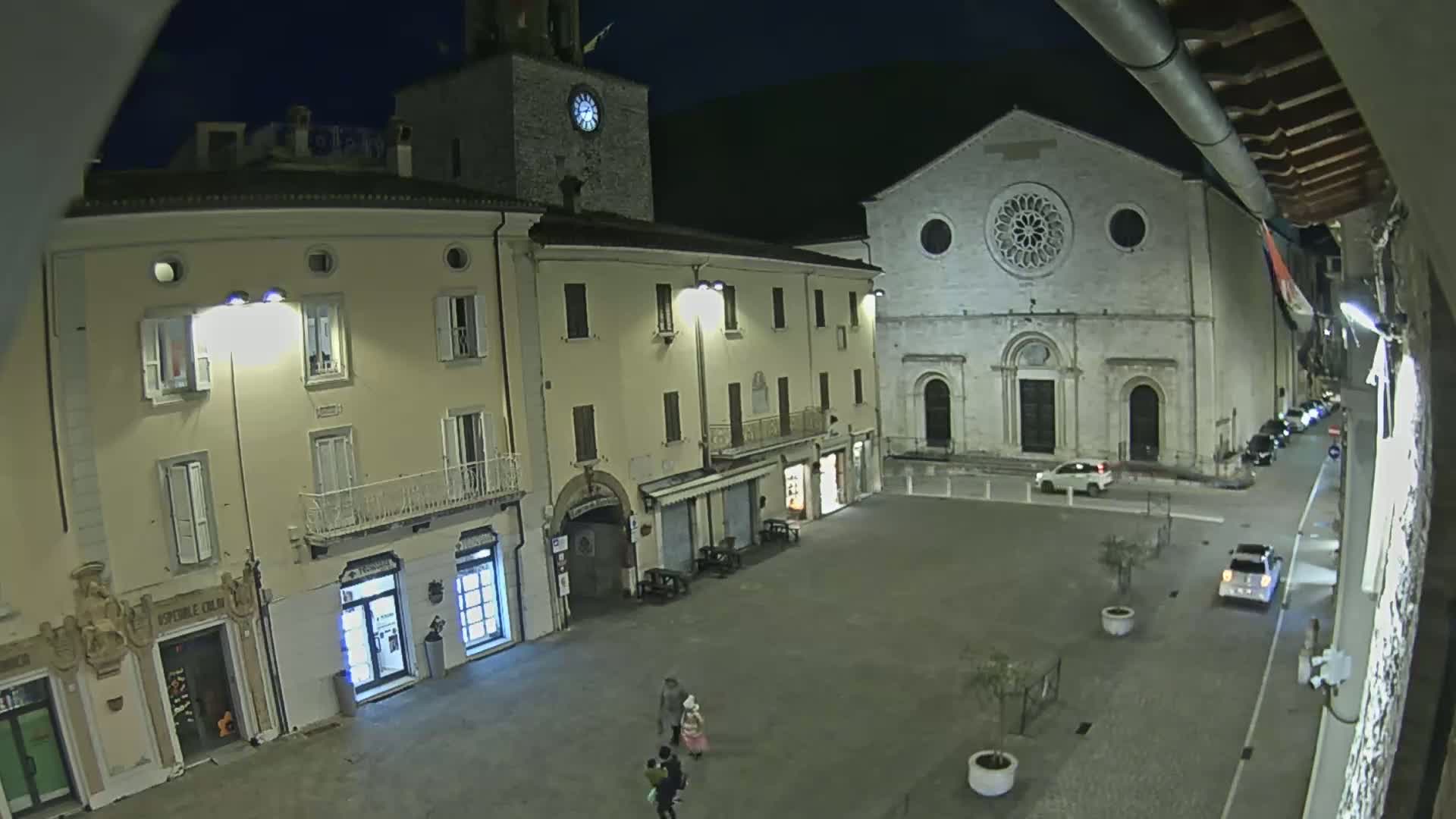 A nighttime view of a town square, featuring several buildings, including a church with a rose window, and a few cars parked along the sides.