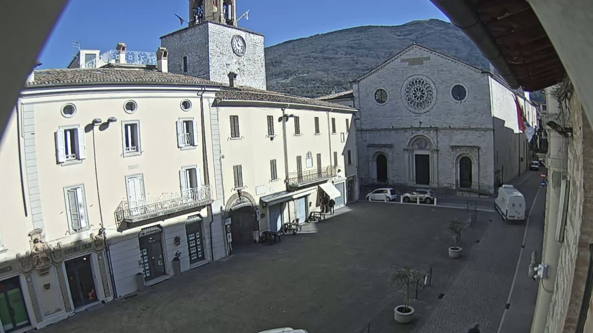 A sunny view of a town square with several buildings, including a church with a rose window and a clock tower, under a clear sky and mountains in the background.