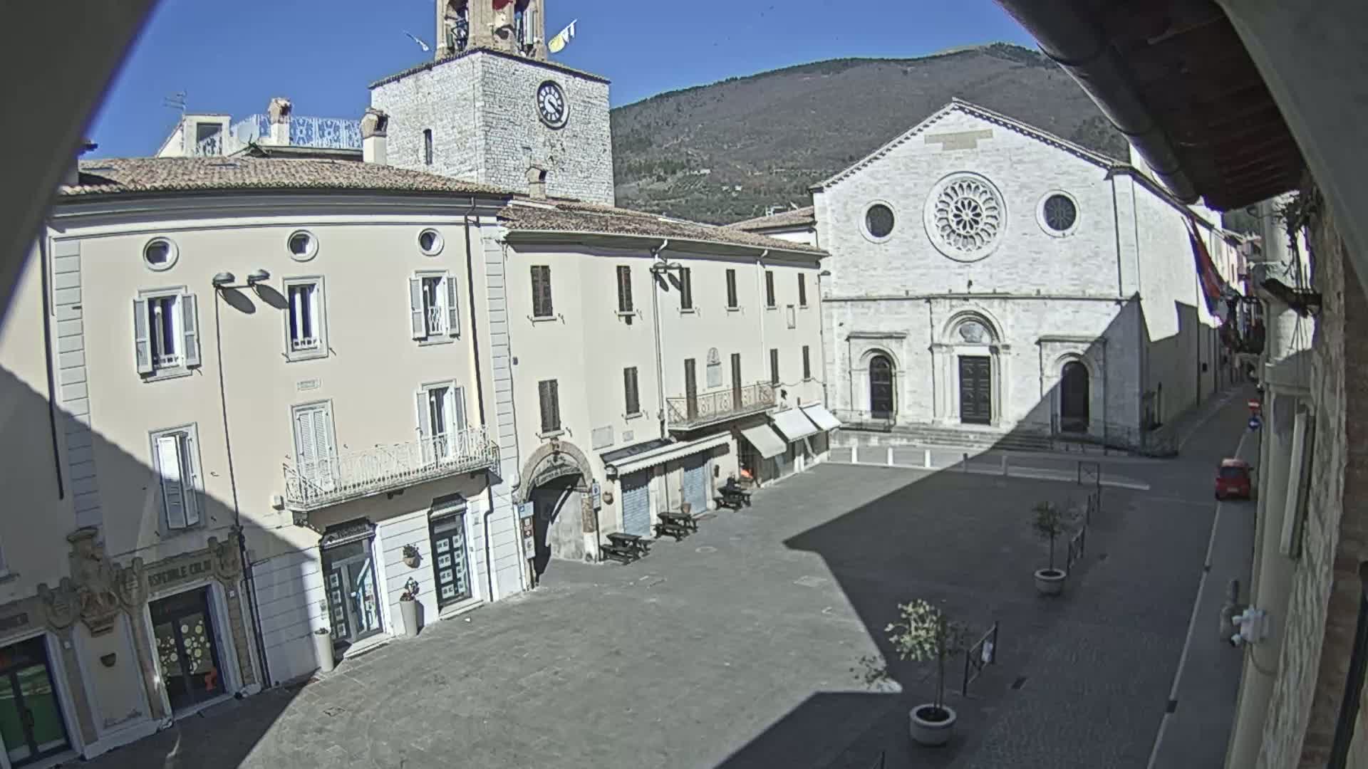 A sunny day reveals a town square with light-colored buildings, including a church with a rose window, and a clock tower, all situated against a backdrop of mountains.