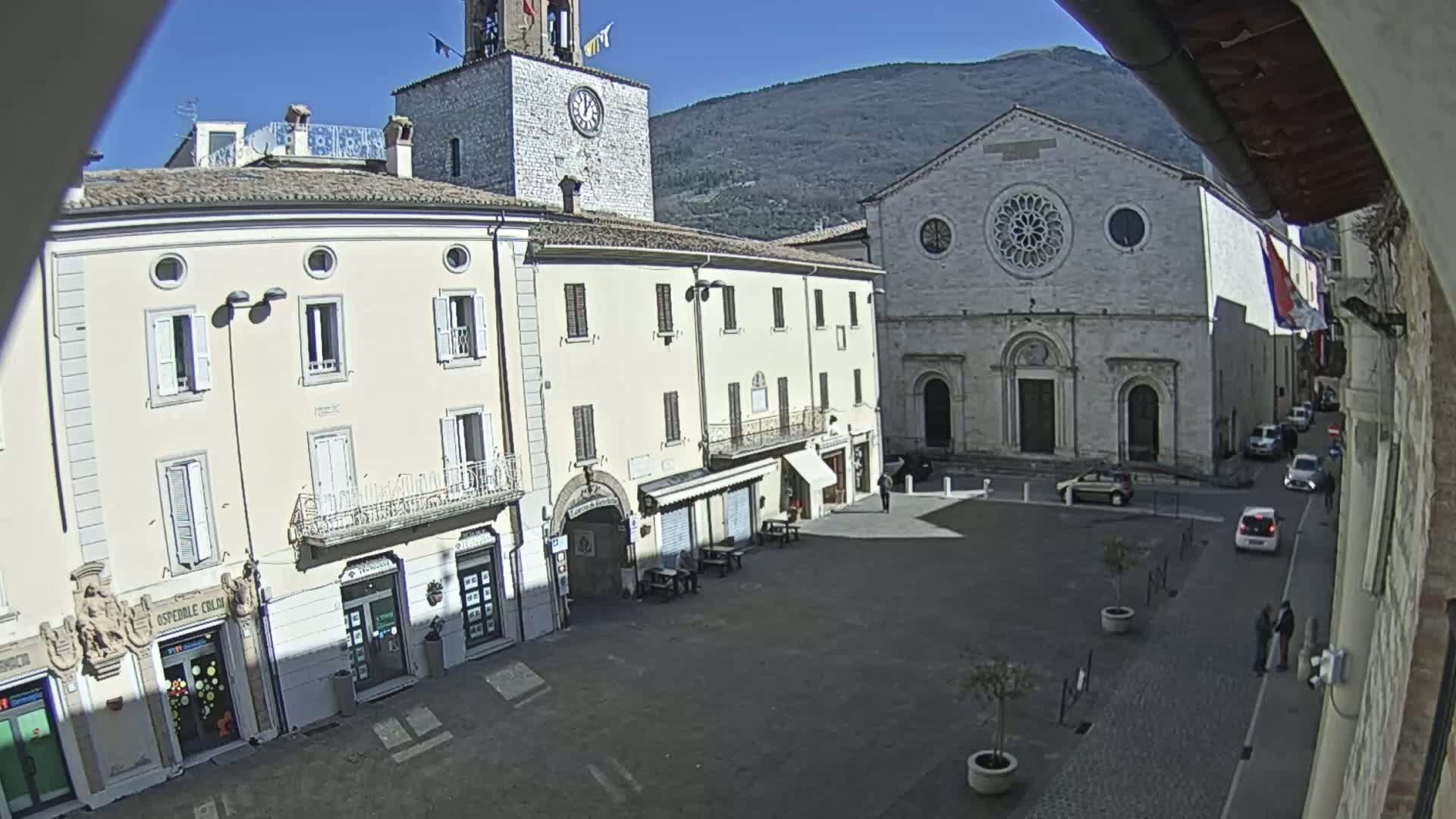 A sunlit town square features several buildings, including a church with a large rose window and a clock tower, under a clear blue sky and mountains in the background.