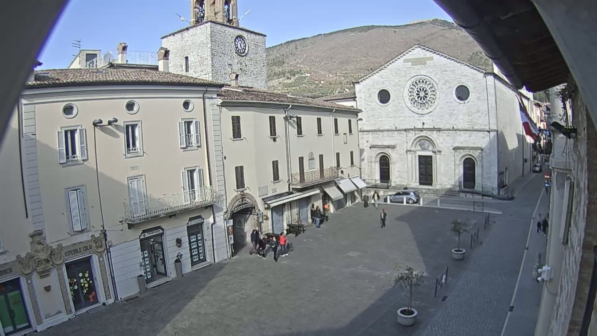 A sunny town square is surrounded by historic buildings, including a church with a rose window and a clock tower, under a clear sky with mountains in the background.