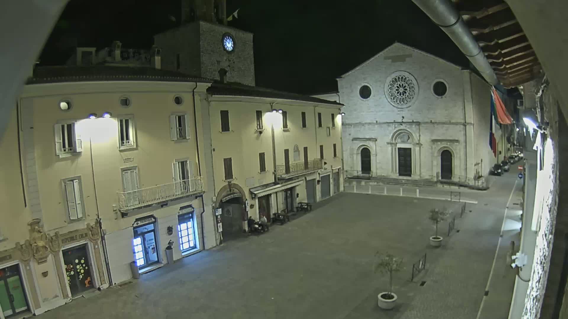 A nighttime view of a town square, illuminated by streetlights, features buildings including a clock tower and a church with a rose window.