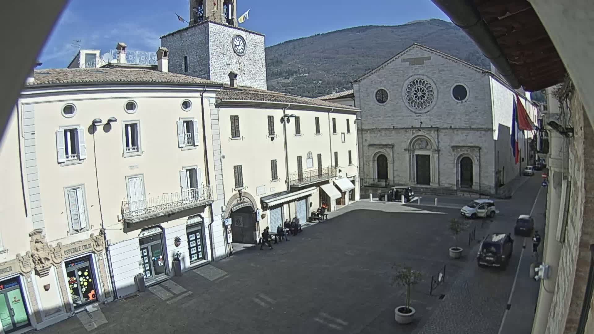 A sunny town square is shown, with several multi-story buildings surrounding it, including a church with a large rose window and a clock tower.