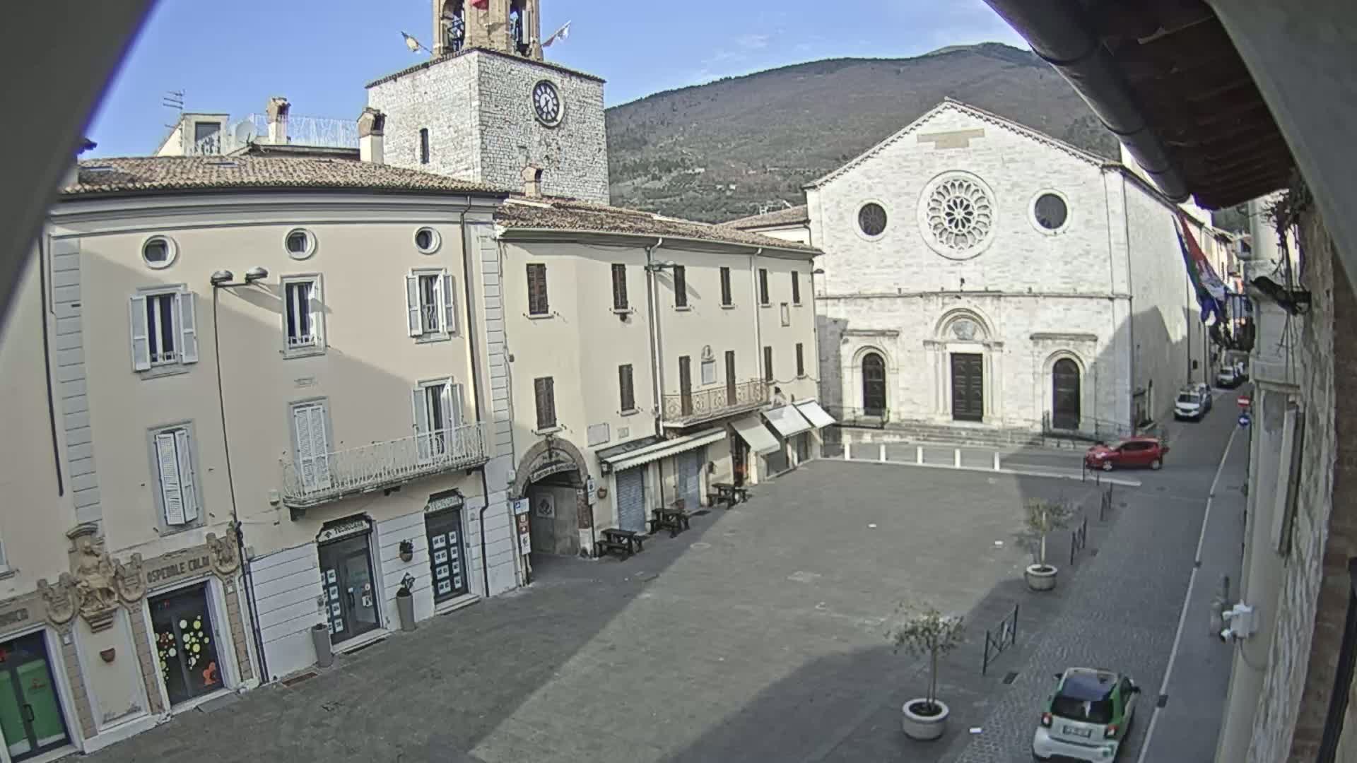 A mostly empty town square is viewed from above on a sunny day, with several buildings surrounding it, including a church with a large rose window and a clock tower.