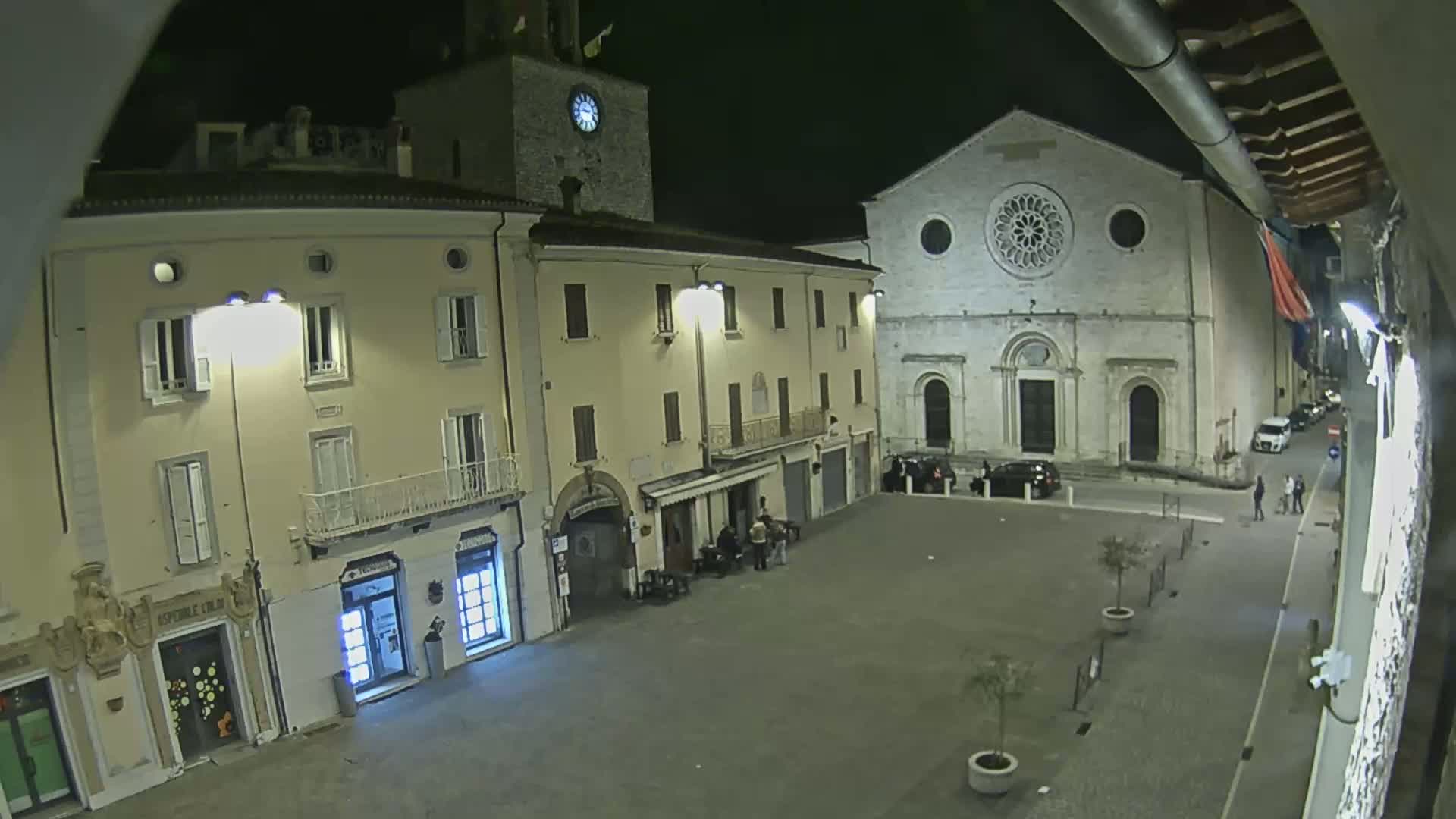A nighttime, high-angle view of a town square with several buildings, including a church with a rose window, and a few people standing around.