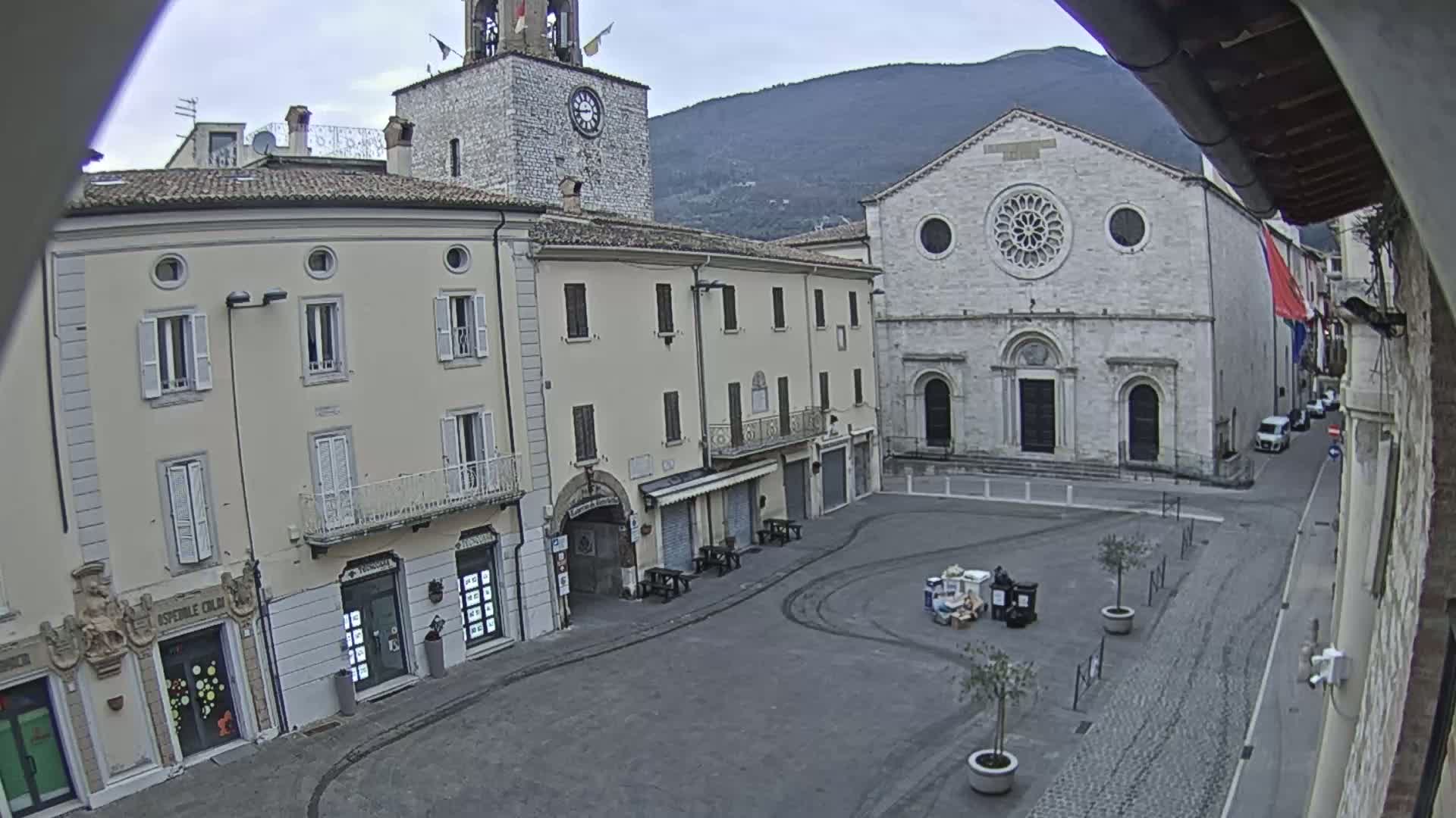 A mostly empty town square is seen on an overcast day, with buildings lining the square including a clock tower and a large stone church, and a street extending from the square.