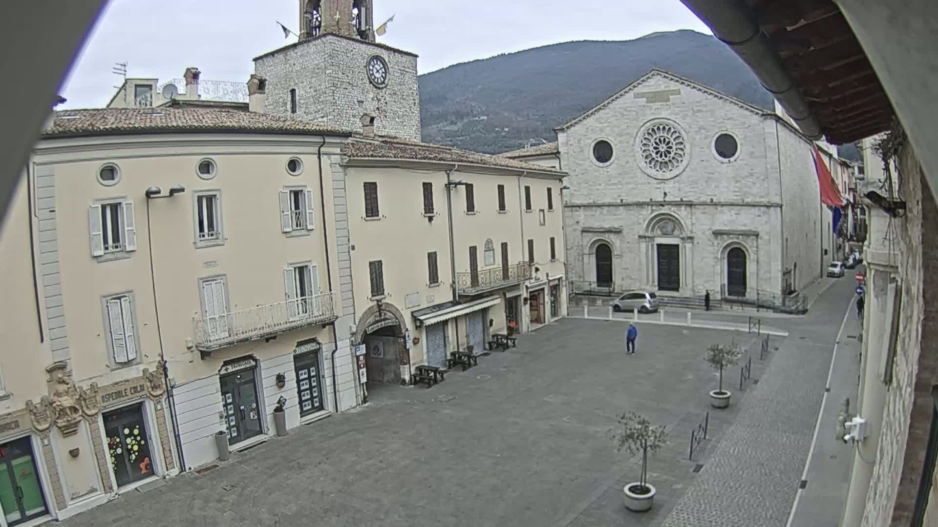 A town square with several light-colored buildings, including a church with a rose window, under an overcast sky.