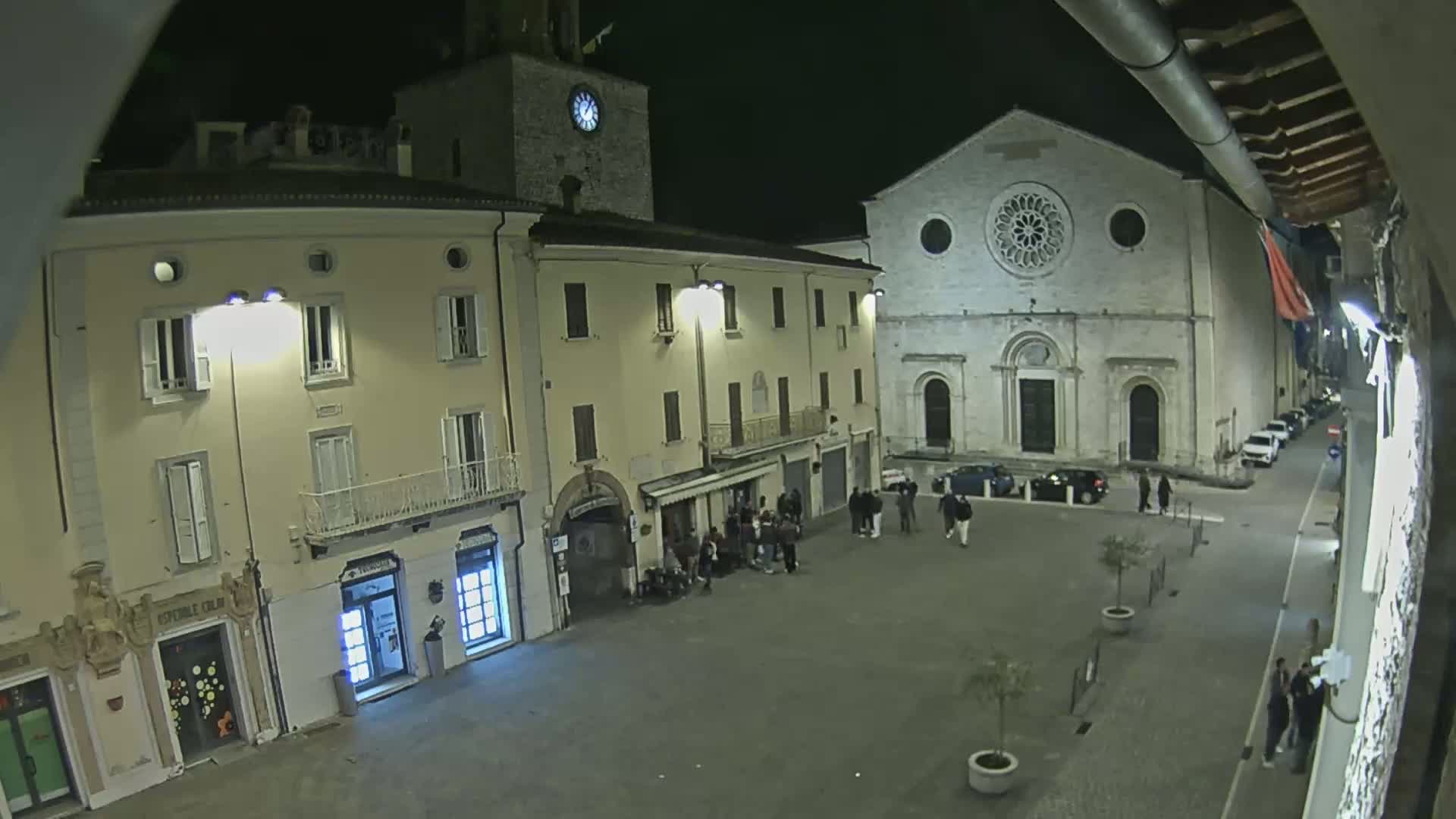 A nighttime view of a town square with several buildings, including a church with a rose window, and a small gathering of people.