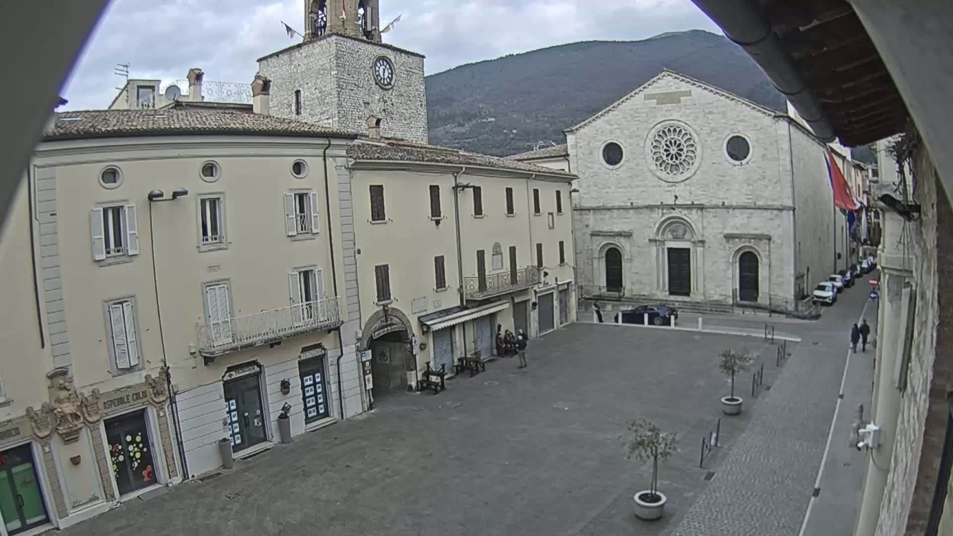 A town square with several light-colored buildings, including a church with a rose window, under an overcast sky.