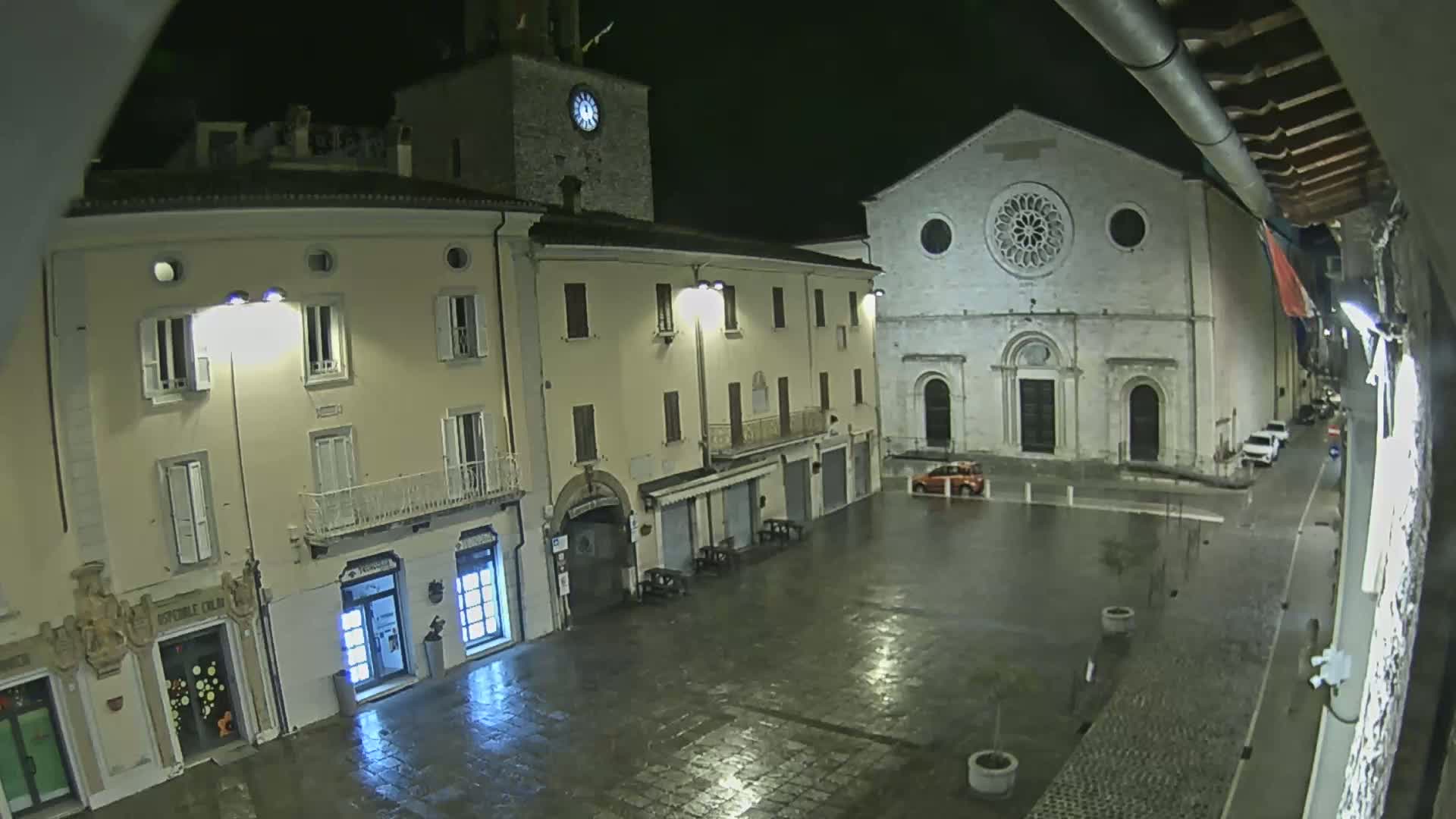 A rain-slicked town square at night is illuminated by streetlights, showing a clock tower, a large church, and several buildings.