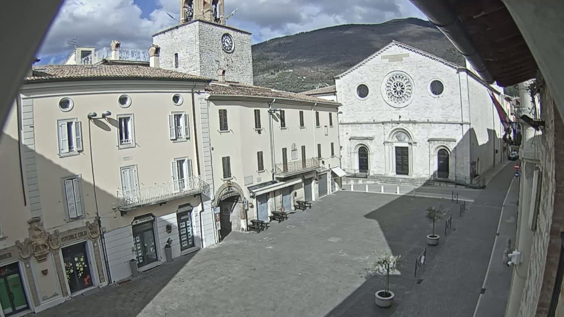 A mostly empty town square with light-colored buildings, including a church with a rose window, under a partly cloudy sky.