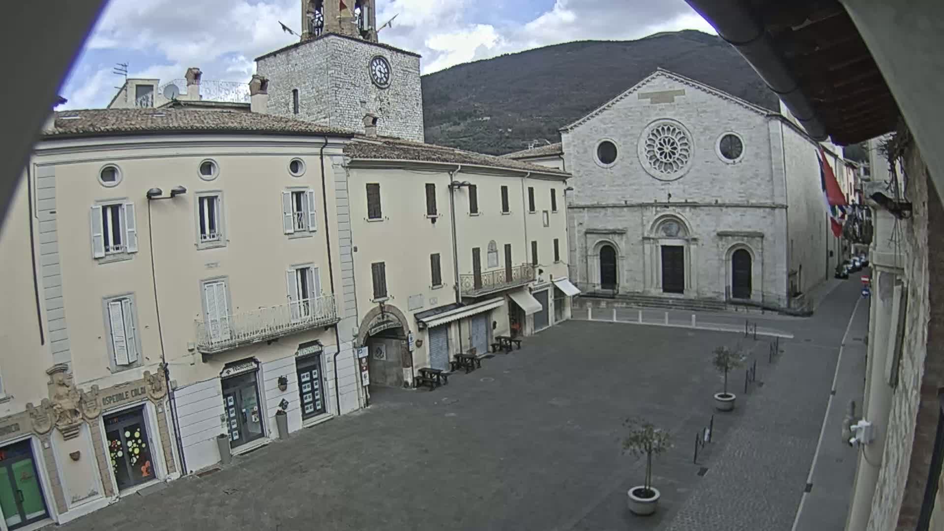 A mostly empty town square is flanked by light-colored buildings, including a church with a rose window, under a cloudy sky.