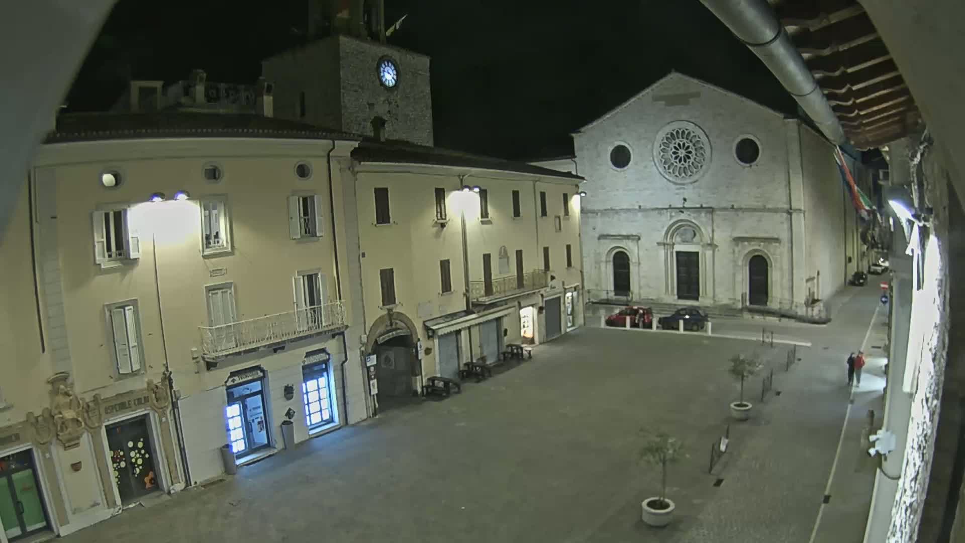 A nighttime view of a town square with several buildings, including a church with a rose window and a clock tower, illuminated by streetlights.