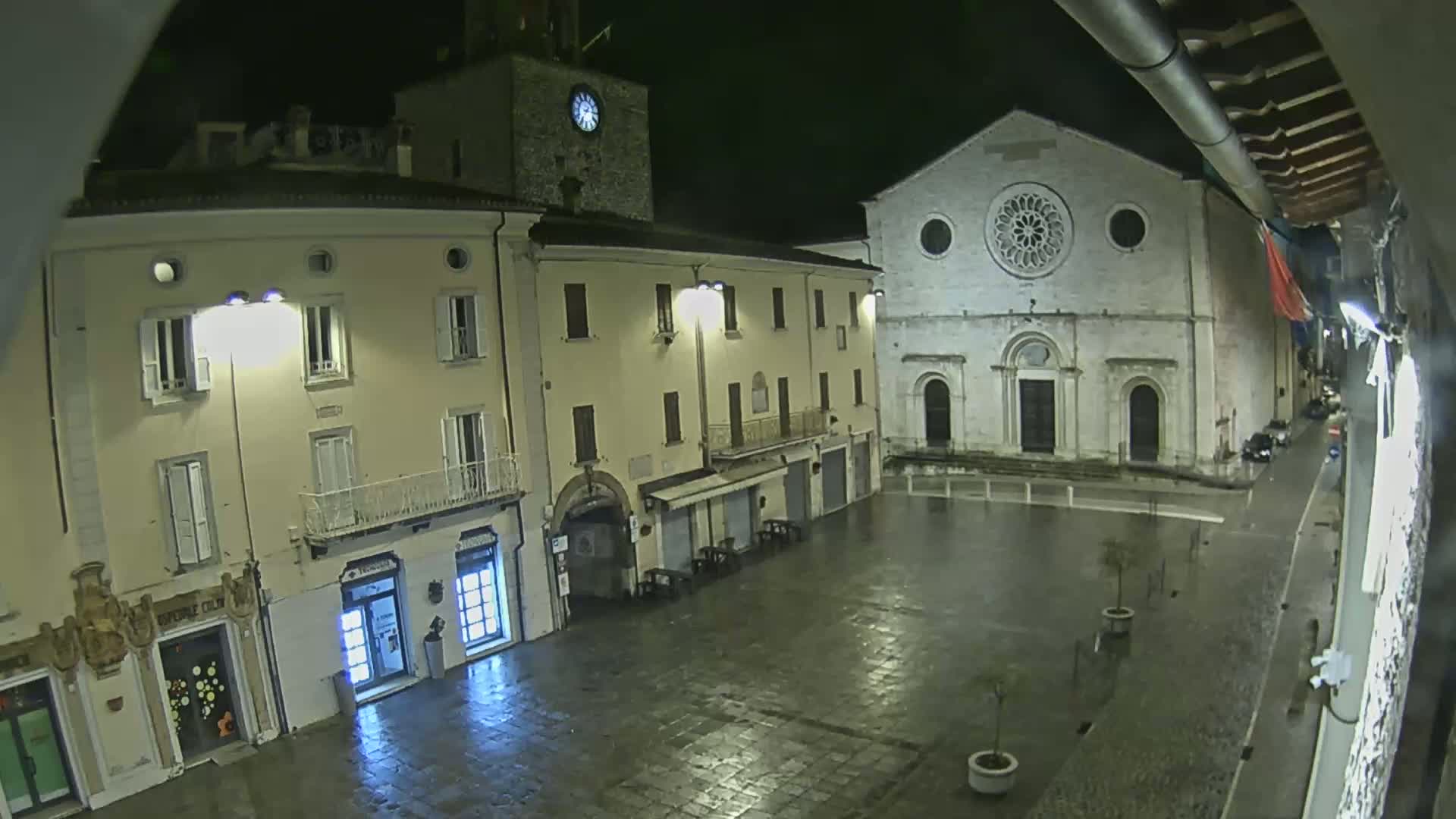 A nighttime view of a town square, wet from rain, showing illuminated buildings including a clock tower and a church with a rose window.