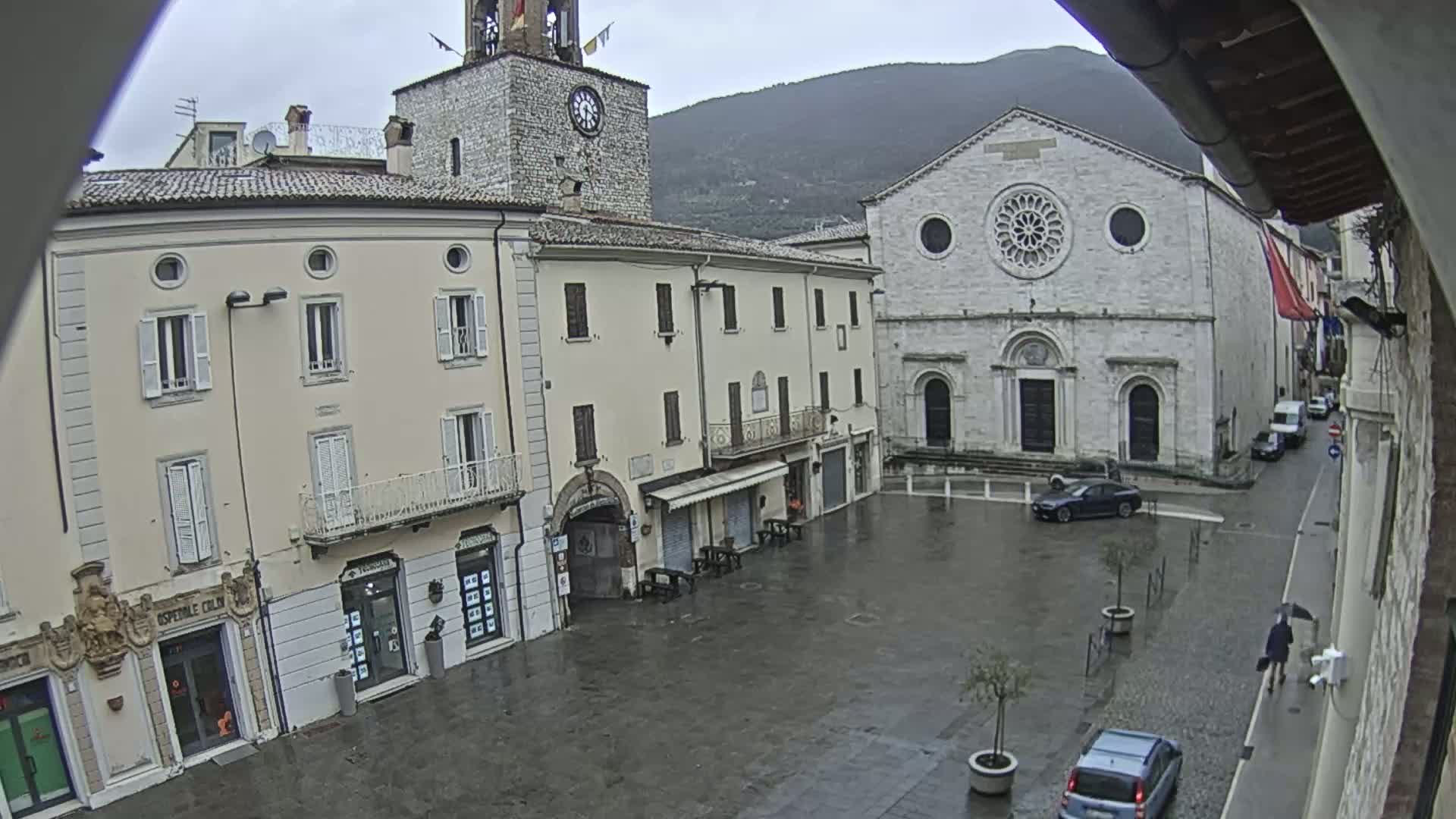A rain-slicked town square is surrounded by historic buildings, including a clock tower and a large stone church, under an overcast sky.