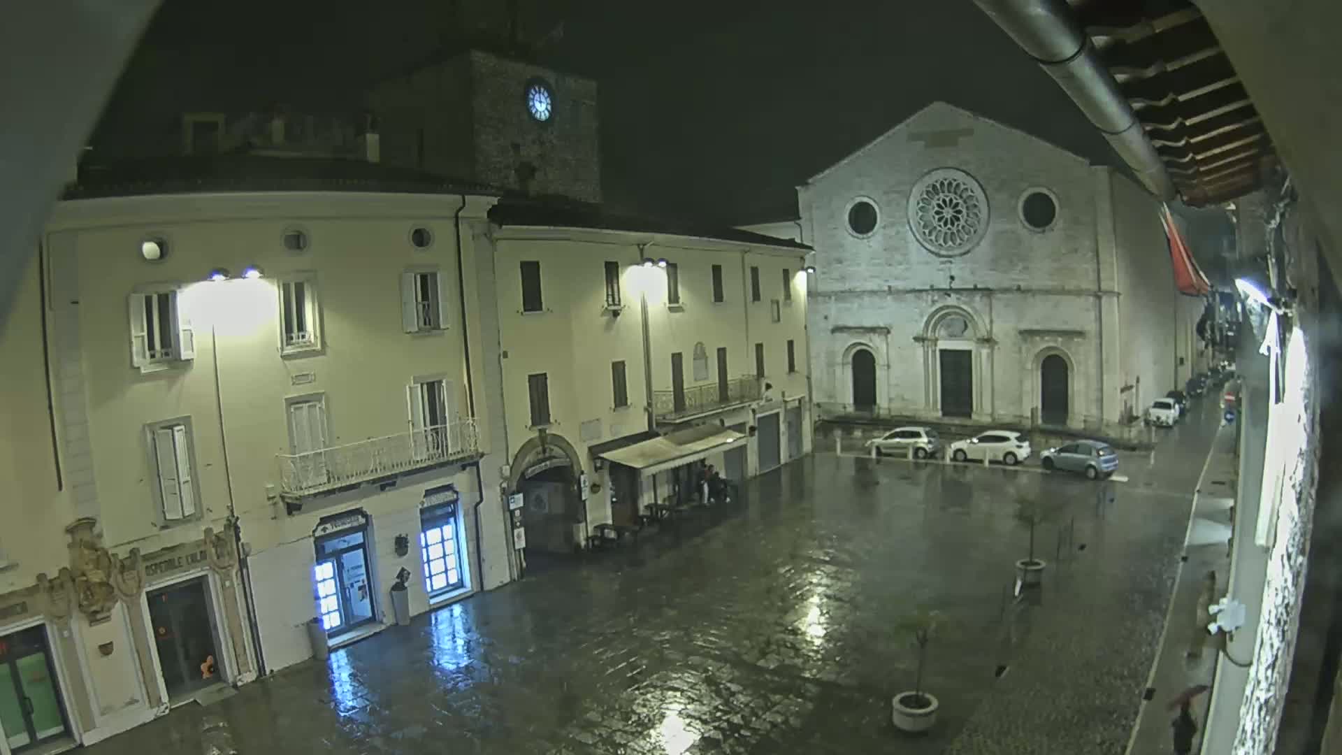 A rain-slicked town square at night is illuminated by streetlights, showing several buildings, including a church with a rose window, and a few parked cars.