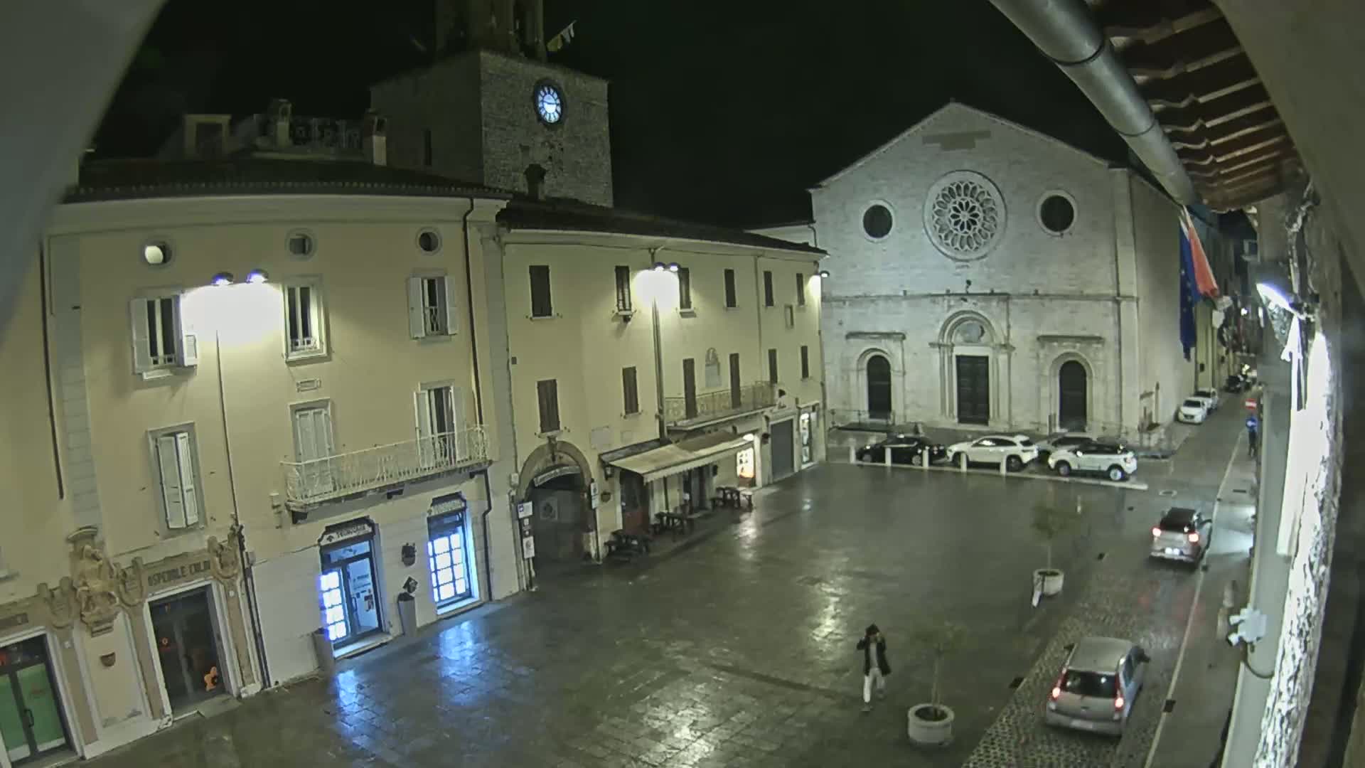 A wet, nighttime town square is shown, featuring several buildings, including a church with a rose window, and a clock tower, with a few cars parked and a lone pedestrian.