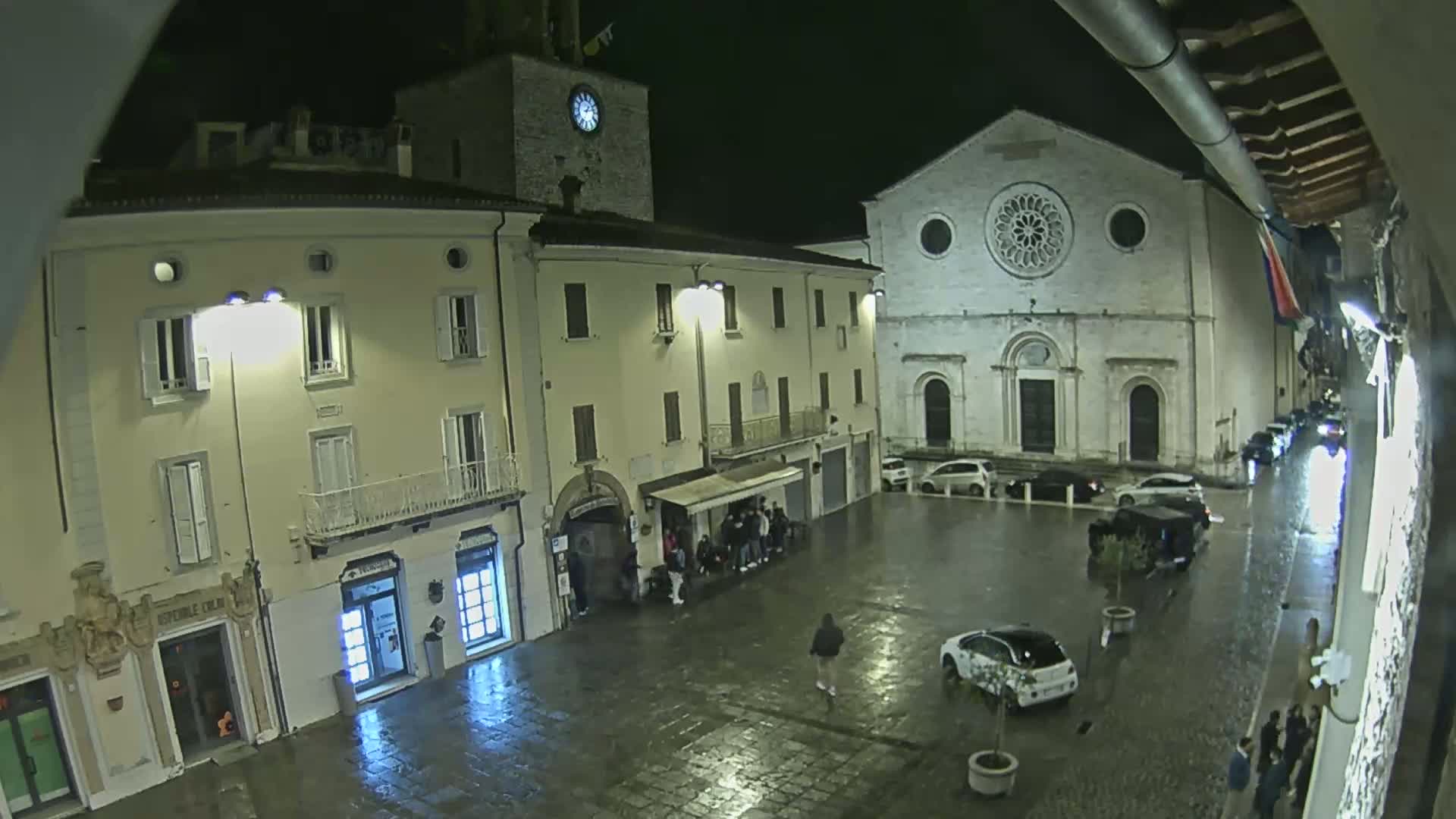 A wet, nighttime town square is shown, featuring several buildings, including a church with a rose window, and a few cars and pedestrians.