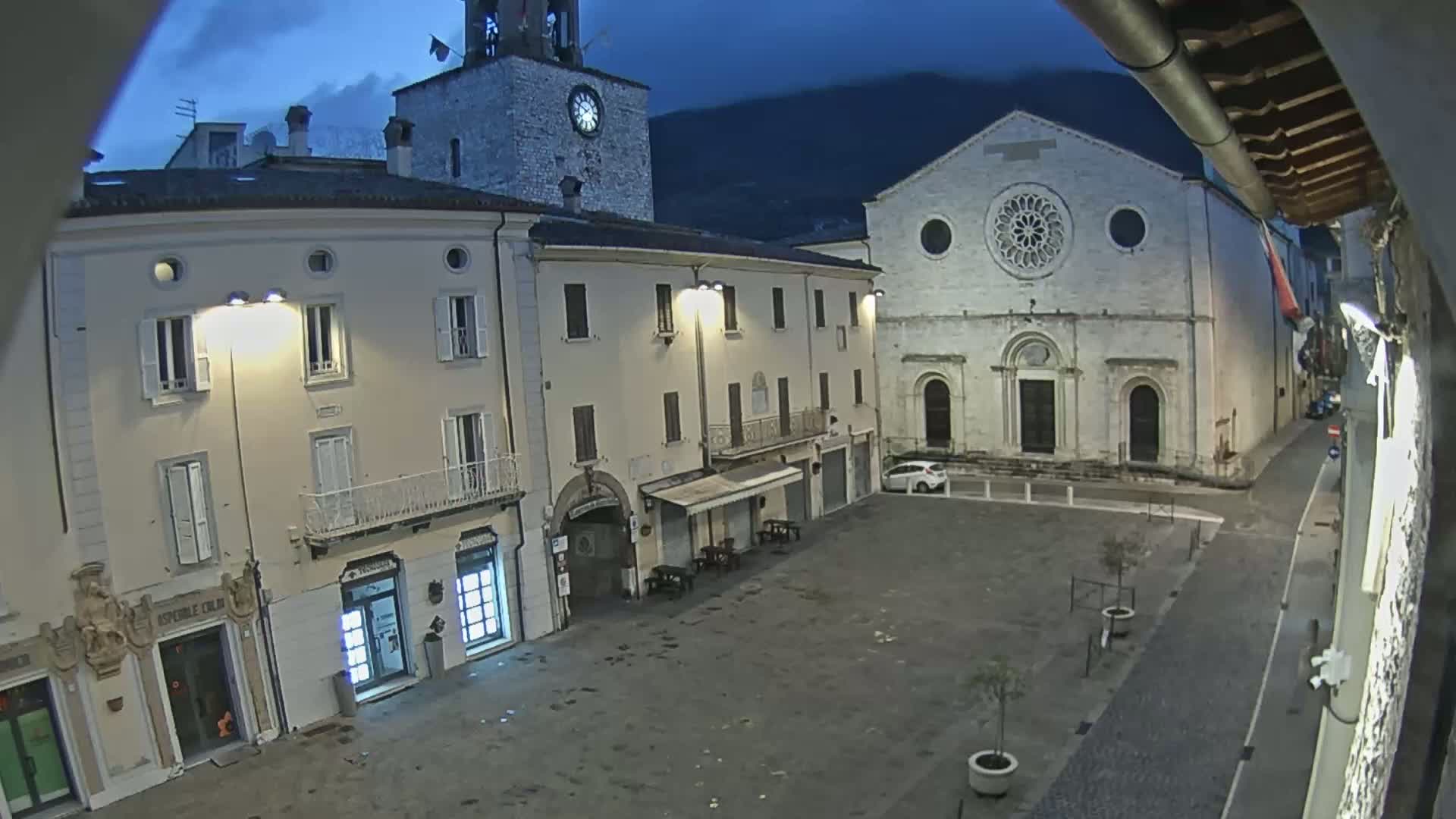A mostly empty town square at night, under a dark sky, is flanked by buildings including a clock tower and a large church.