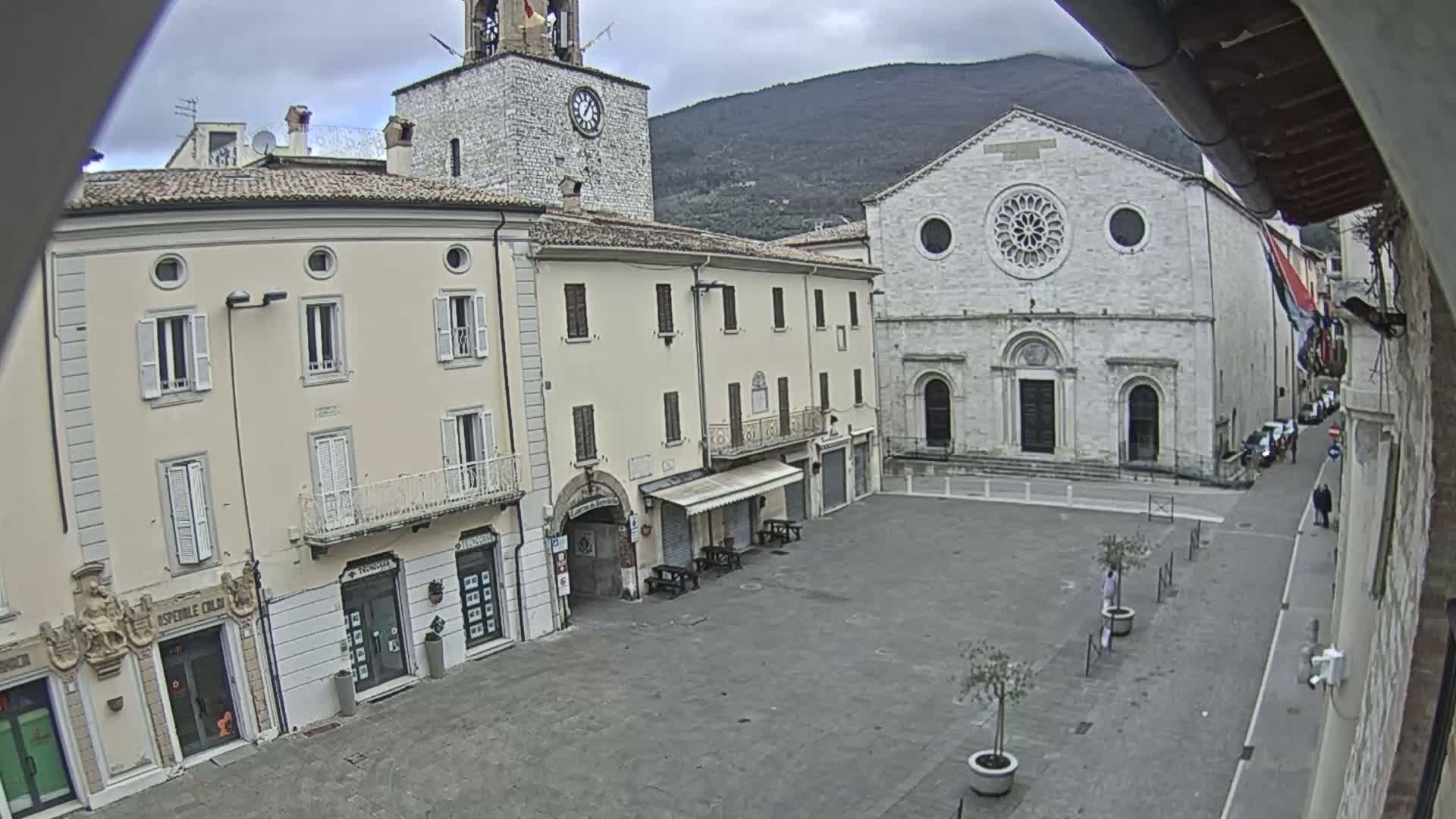 A mostly empty town square is shown on an overcast day, with several light-colored buildings surrounding it, including a clock tower and a church with a large rose window.