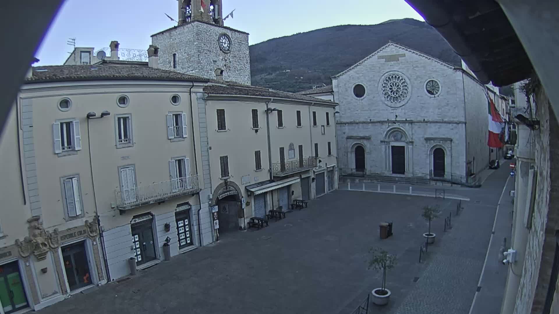 A mostly empty town square is shown under a clear, light sky, with several buildings surrounding it, including a clock tower and a large church.