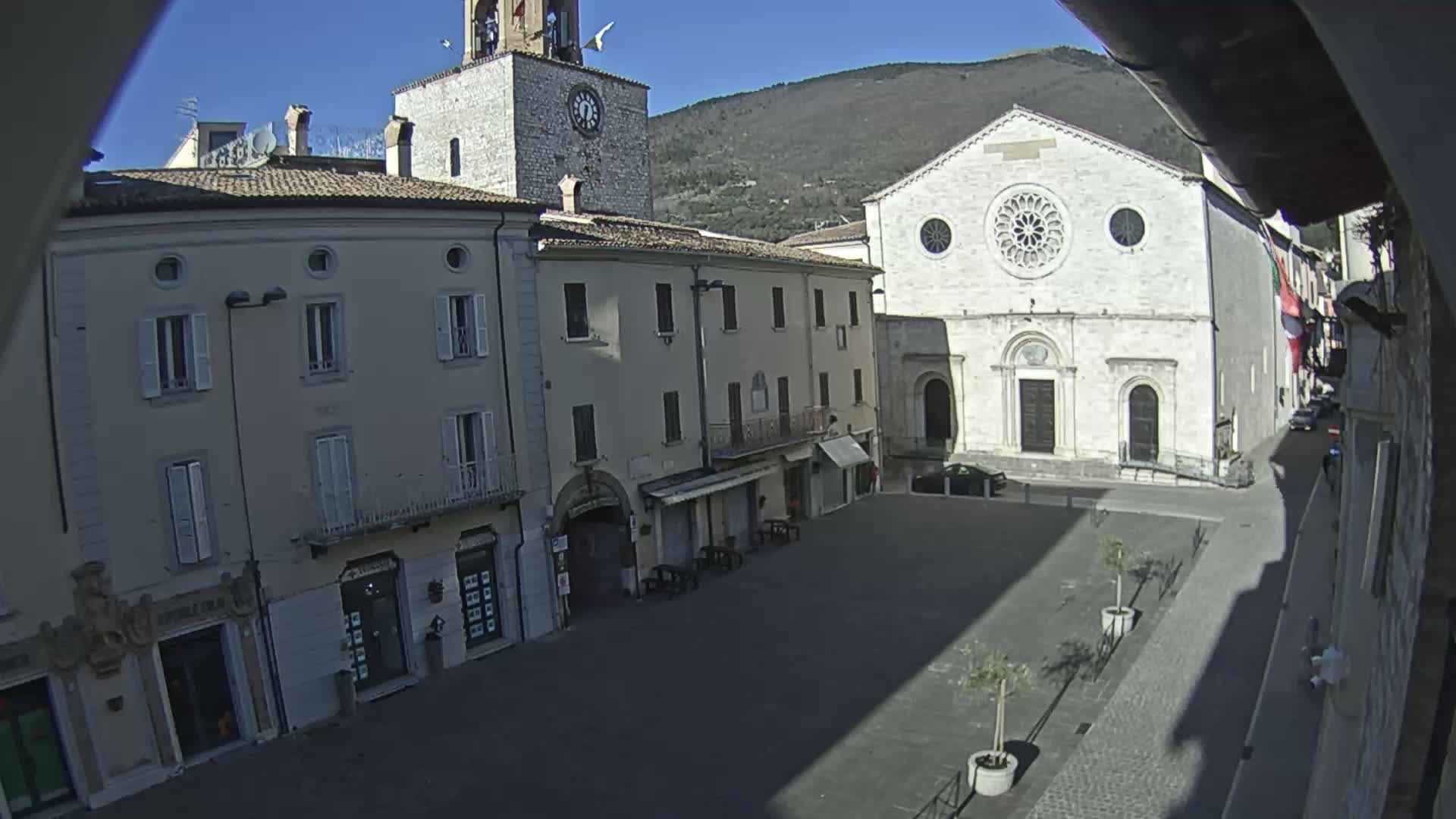 A sunny view of a town square, featuring light-colored buildings, including a church with a rose window, and a clock tower, under a clear blue sky.