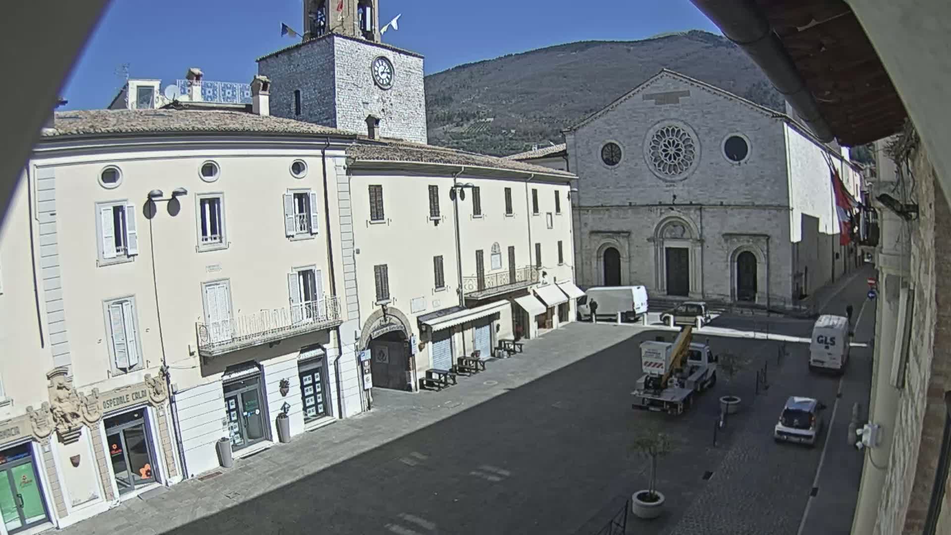A sunny day reveals a town square with several light-colored buildings, including a church with a rose window and a clock tower, and a few vehicles are parked or driving through the square.