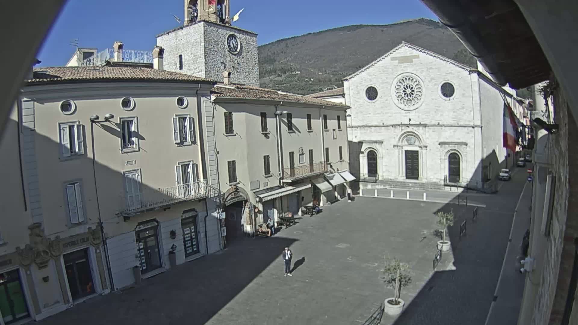 A sunny day reveals a town square with several buildings, including a large church and a clock tower, surrounded by a few people and small trees in planters.