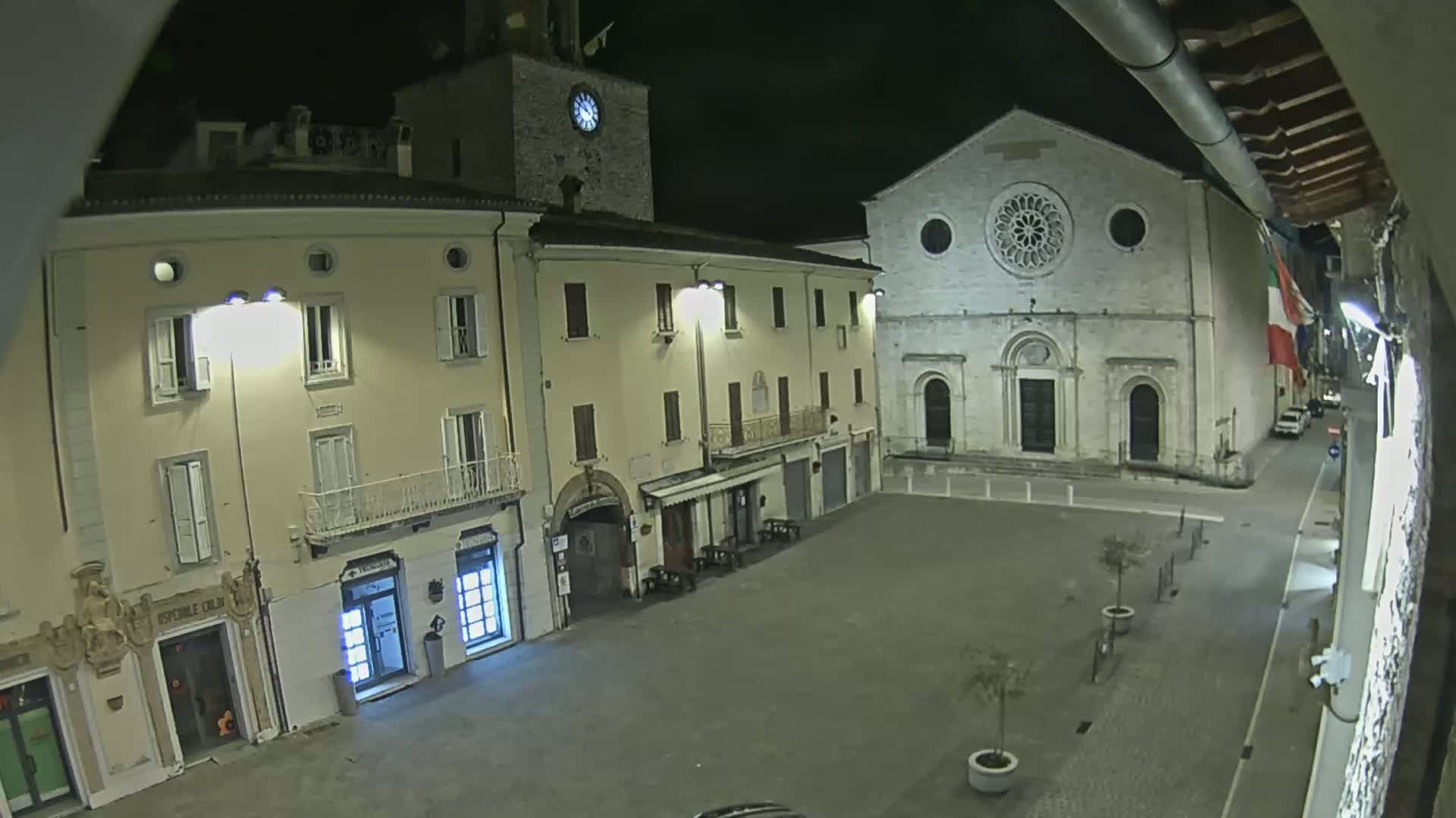 A nighttime view of a town square, featuring several buildings illuminated by streetlights, including a church with a rose window and a clock tower.