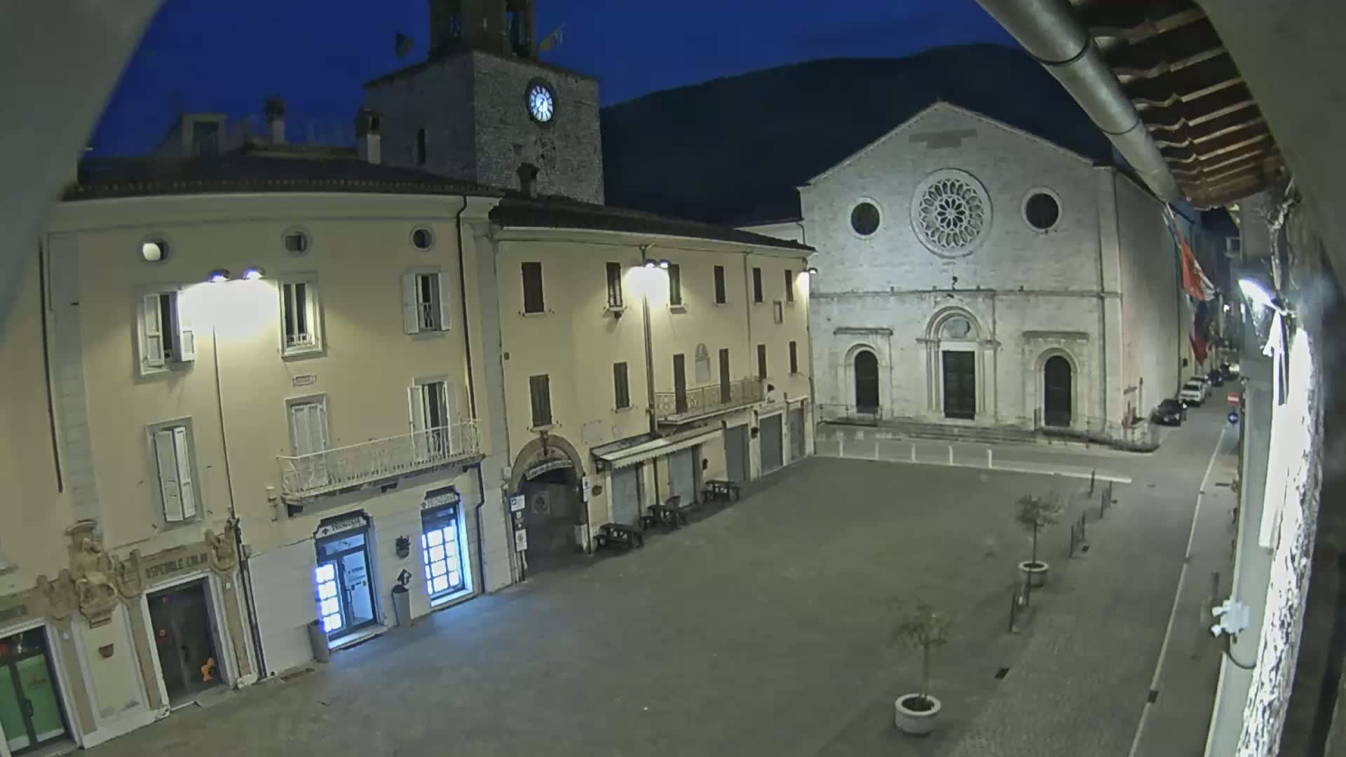 A nighttime view of a town square, featuring a clock tower, a large stone church, and several buildings under illuminated streetlights.