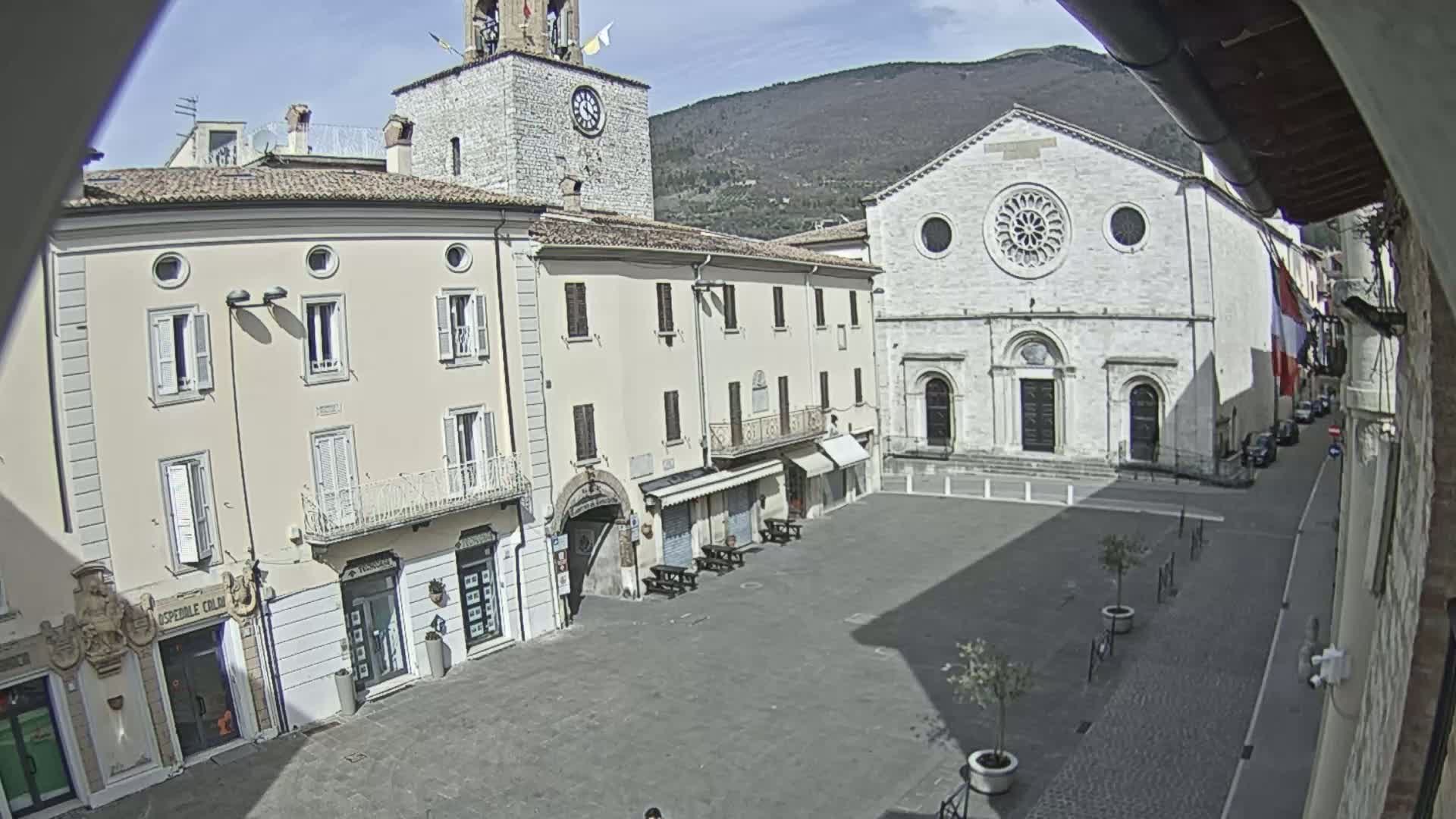 A mostly empty town square is viewed from above on a sunny day, with a large stone church and clock tower visible, and mountains in the background.