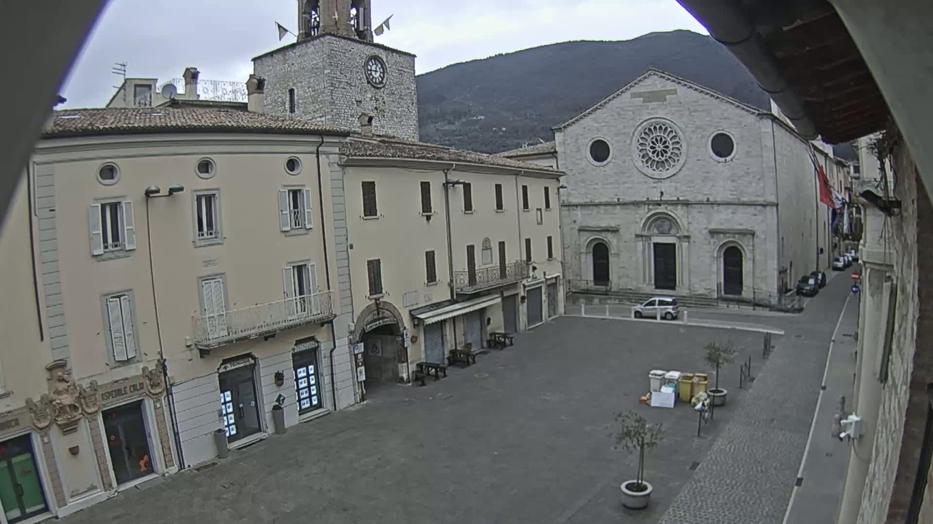 A mostly empty town square is seen on an overcast day, with several multi-story buildings surrounding it, including a church with a large rose window and a clock tower.