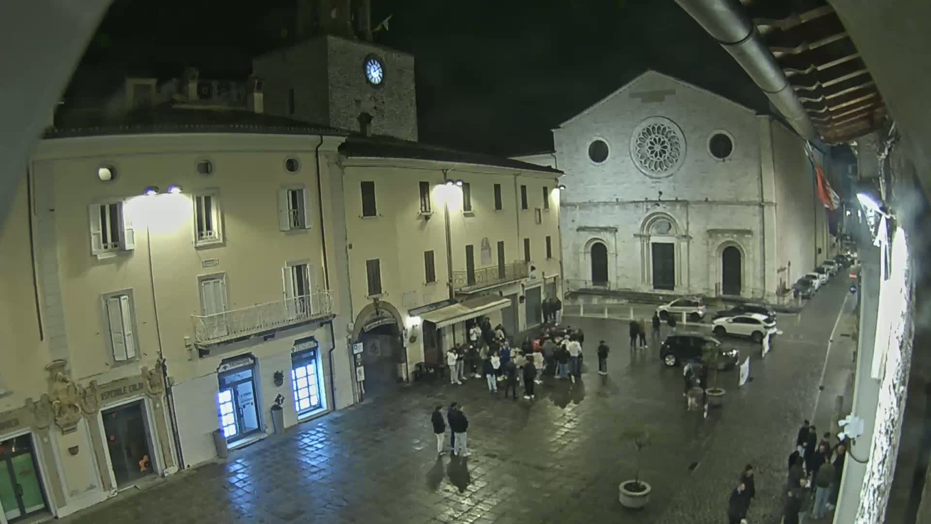 A nighttime, wet town square shows several people gathered near buildings, one of which is a church with a large rose window.