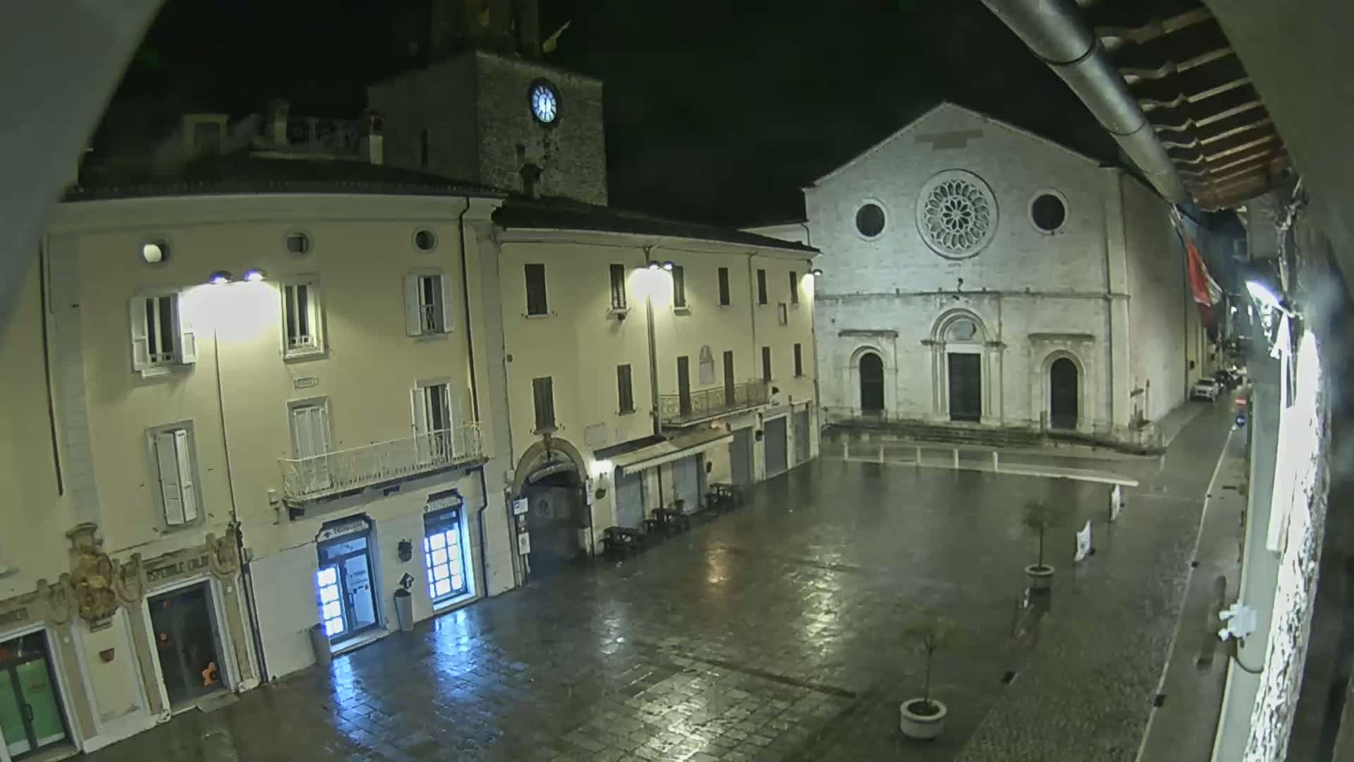 A rain-slicked town square at night is illuminated by streetlights, showing buildings including a church with a rose window and a clock tower.