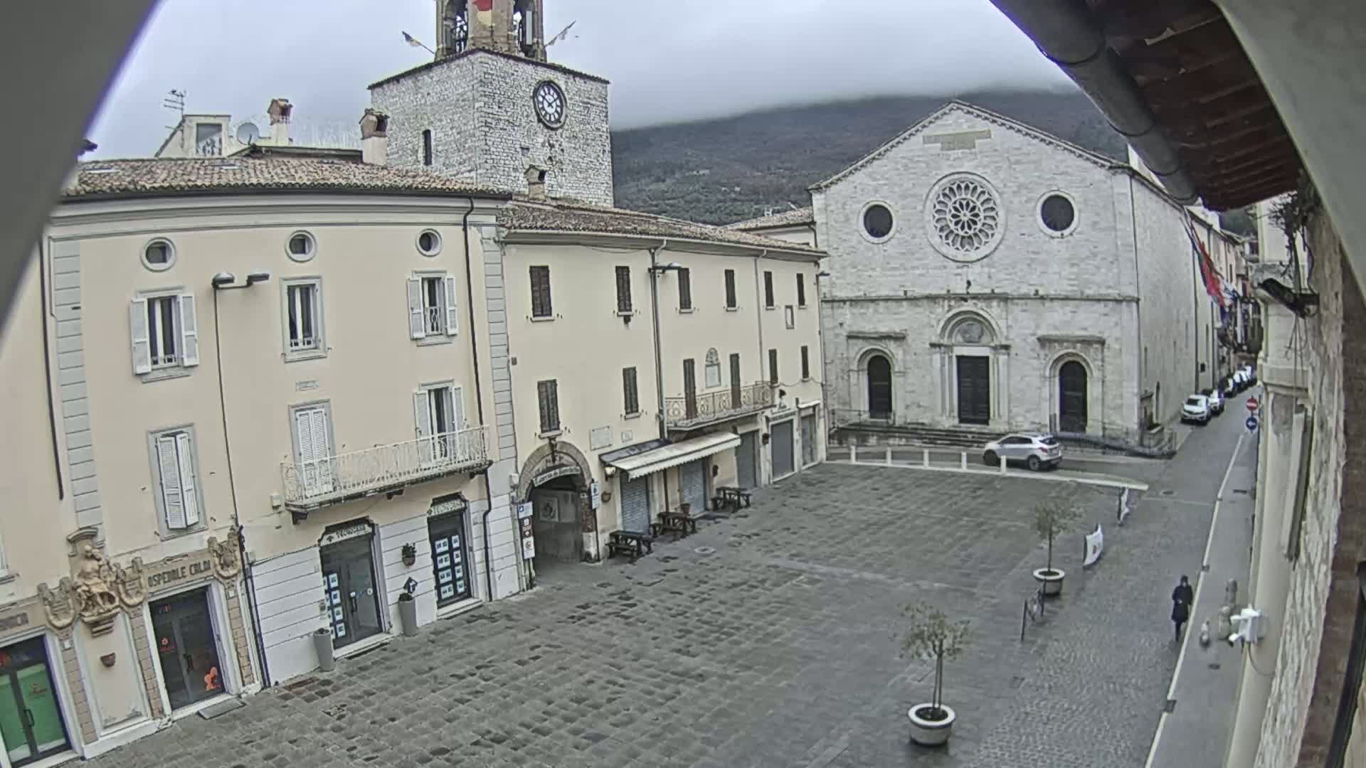 A town square with light-colored buildings, including a church with a rose window and a clock tower, on an overcast day.