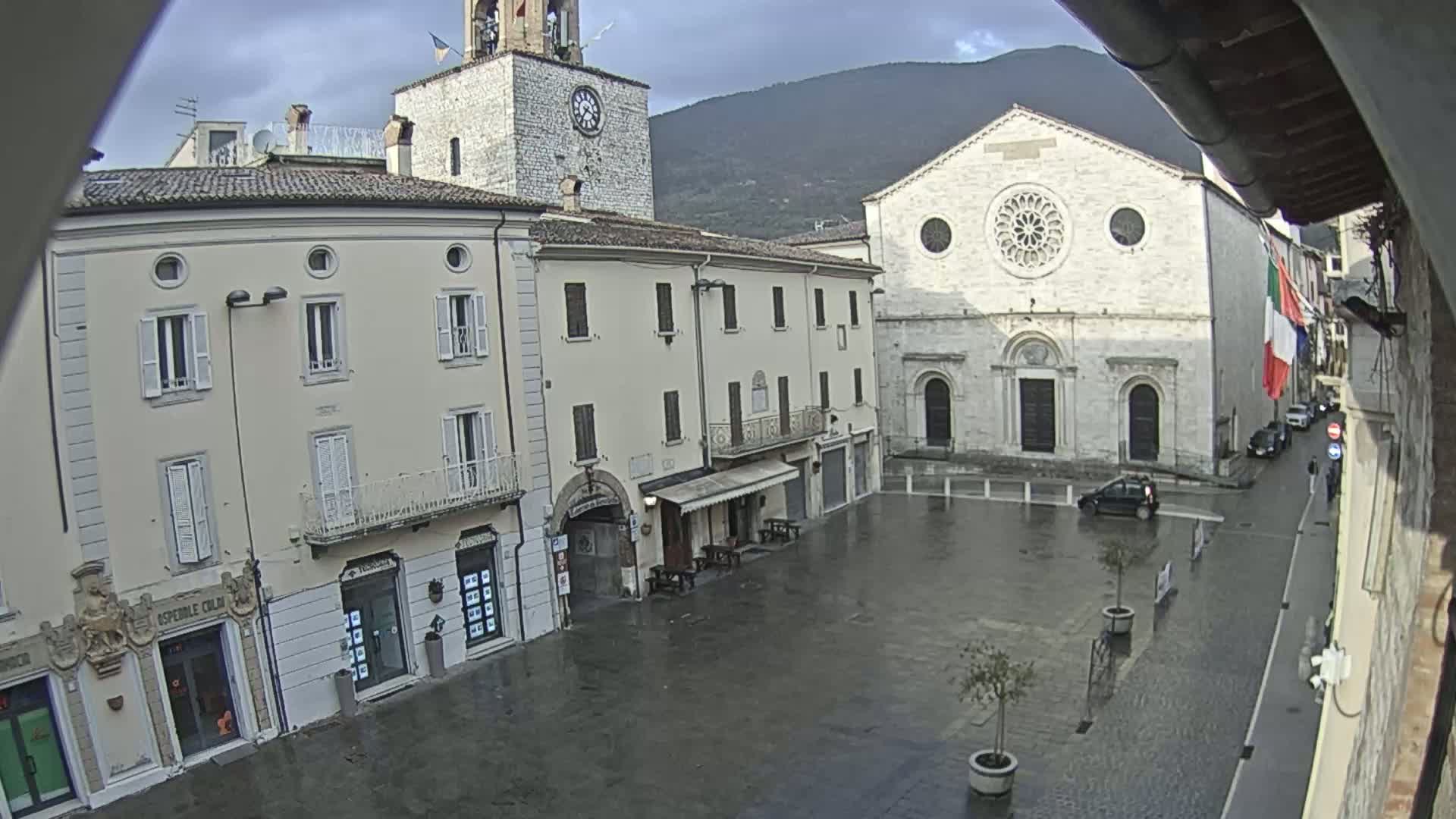 A wet town square is surrounded by several buildings, including a church with a large rose window, under a cloudy sky.