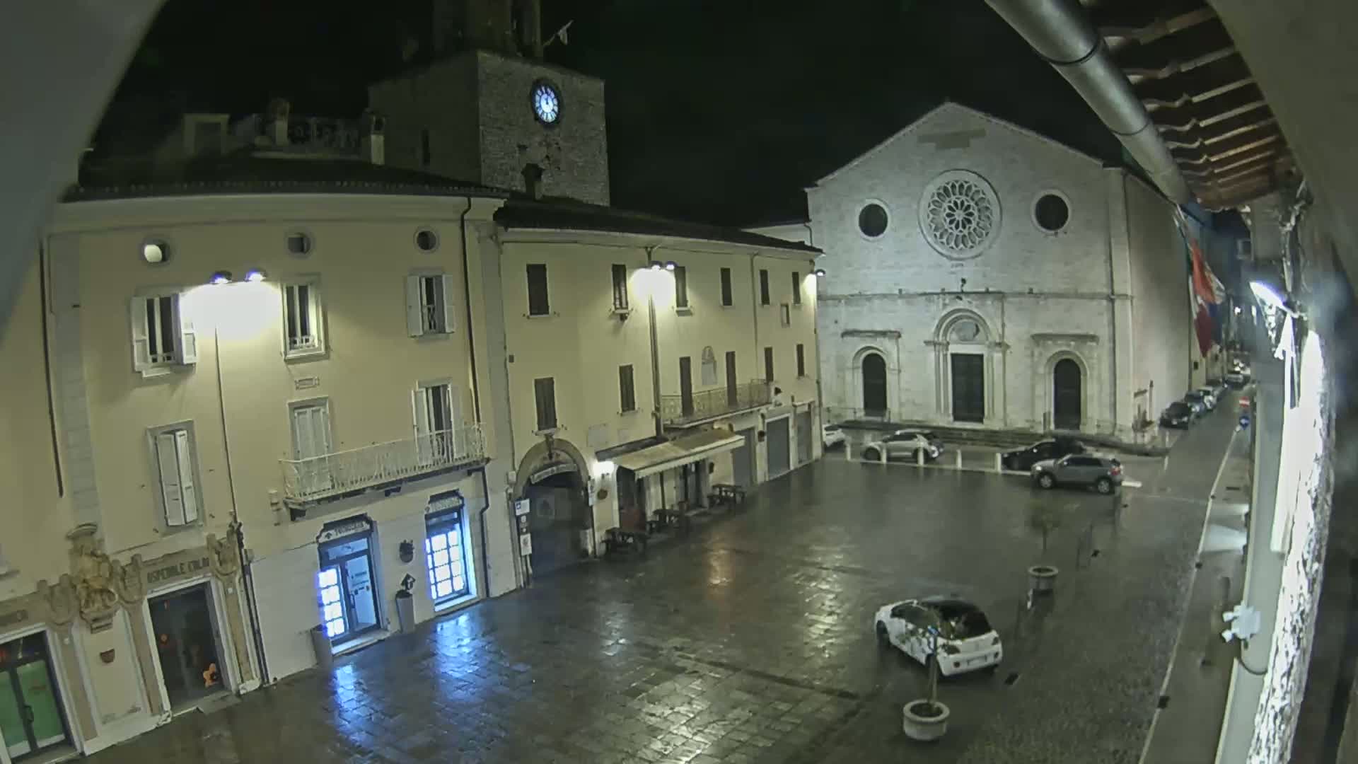 A rain-slicked town square at night, featuring several buildings including a church with a large rose window, illuminated by streetlights.