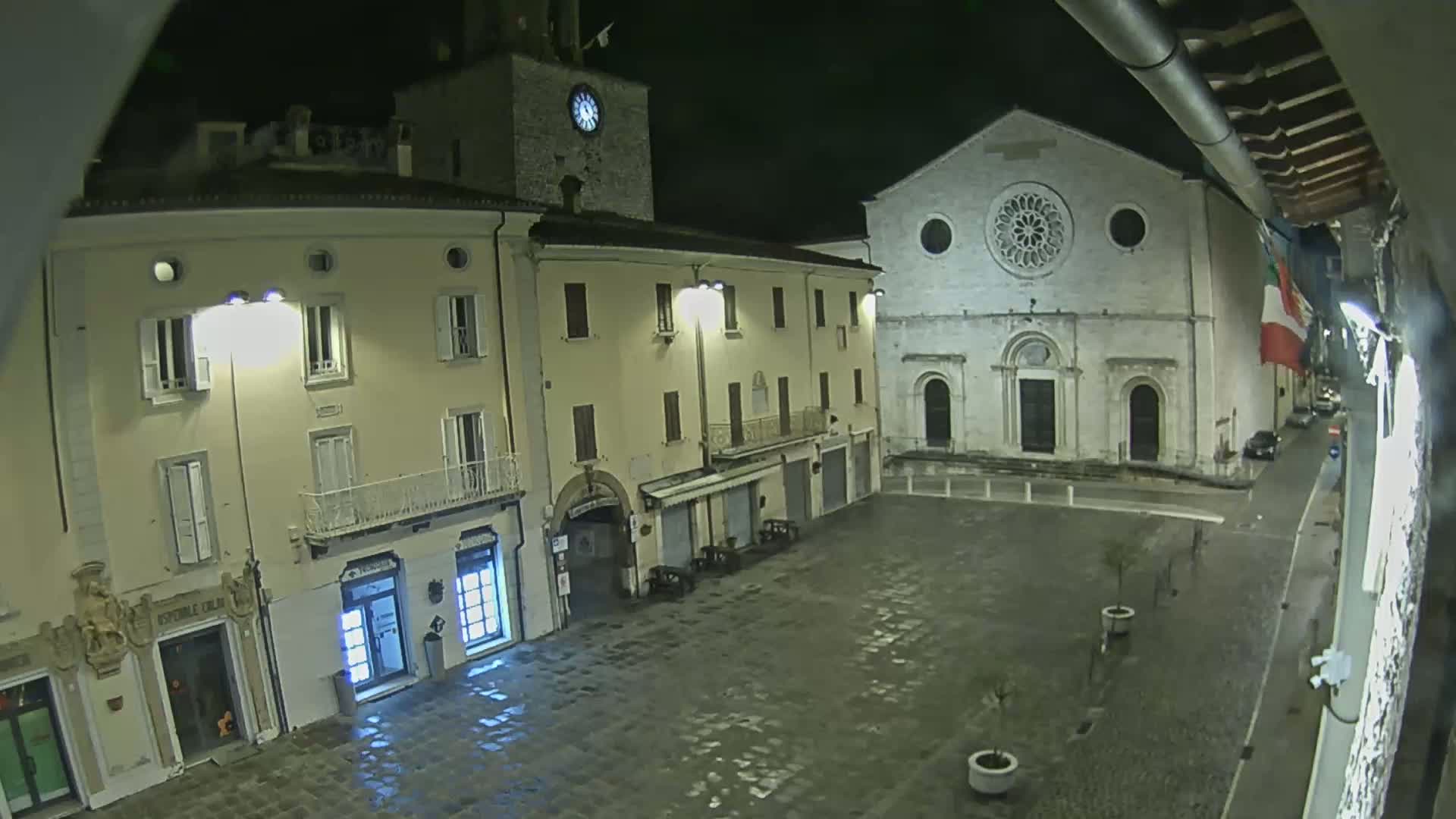 A nighttime view of a town square with buildings, including a church with a rose window, illuminated by streetlights on a wet surface.