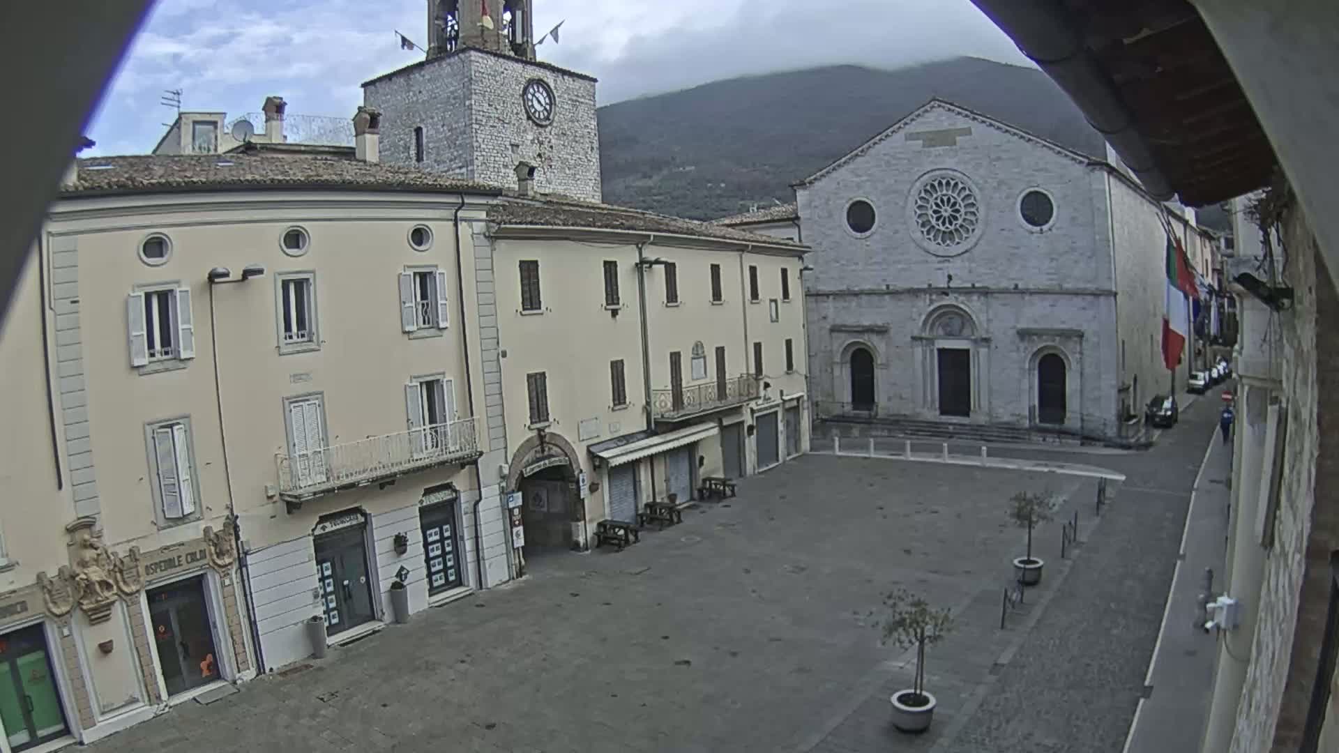 An overcast day reveals a town square with several buildings, including a clock tower and a church with a rose window, nestled against a mountain.