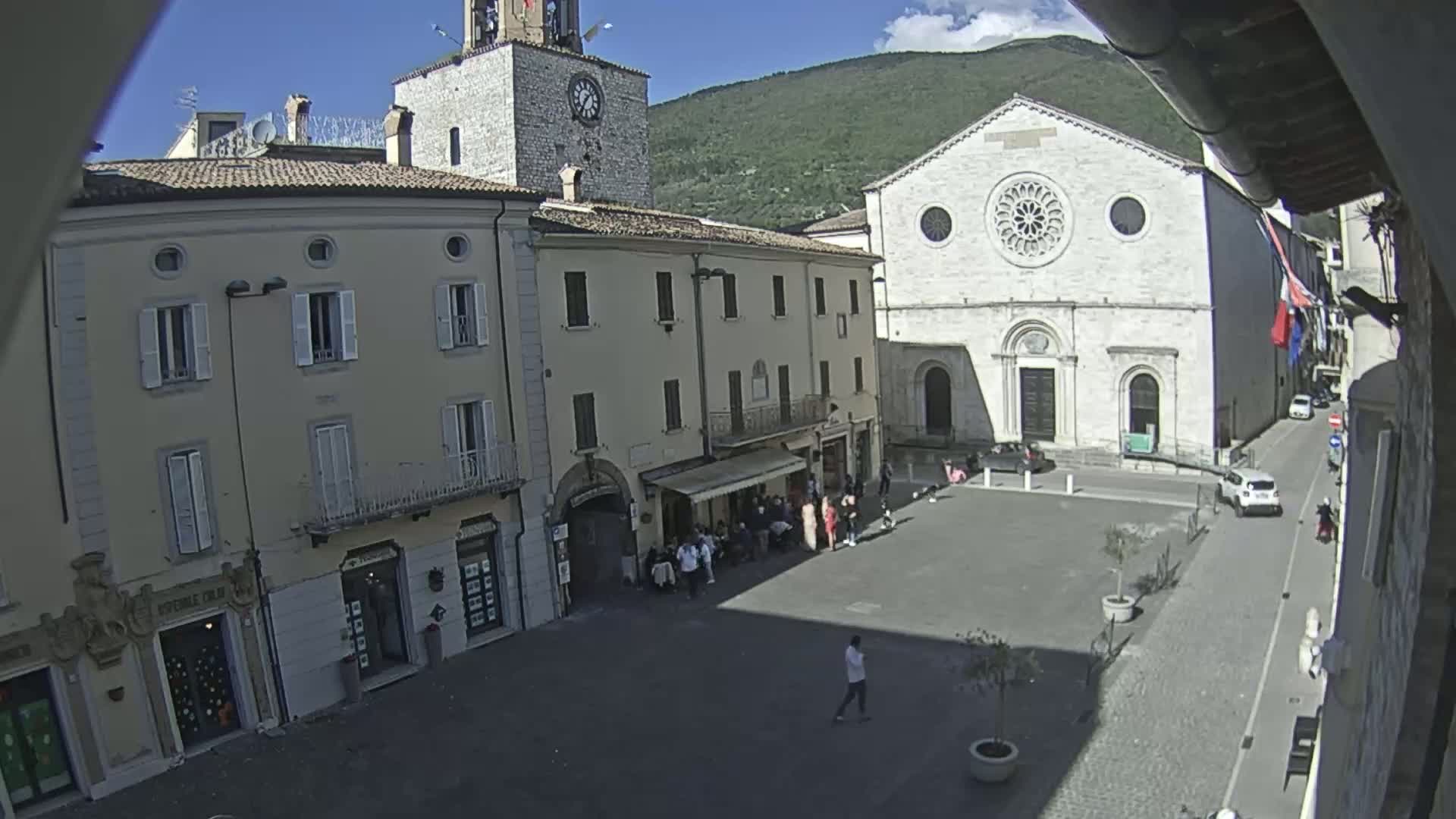 A sunny town square is shown, with several people gathered in front of buildings, including a church with a large rose window and a clock tower.