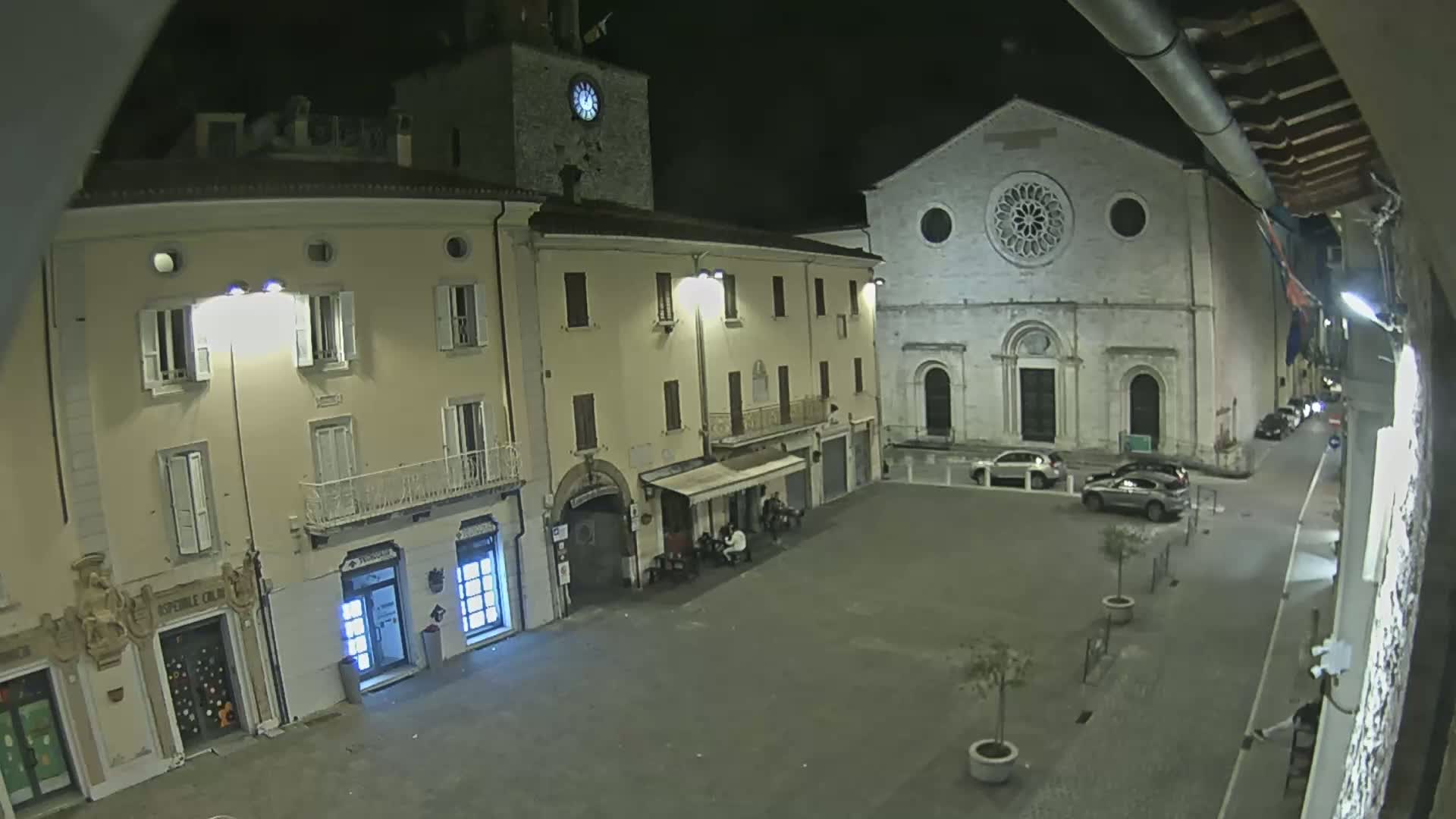 A nighttime view of an empty town square with several buildings, including a church with a rose window, illuminated by streetlights.