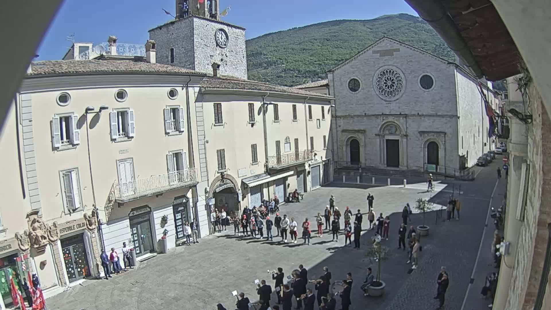 A town square with several buildings, including a church and clock tower, is filled with people standing in groups and a brass band playing under a sunny sky.