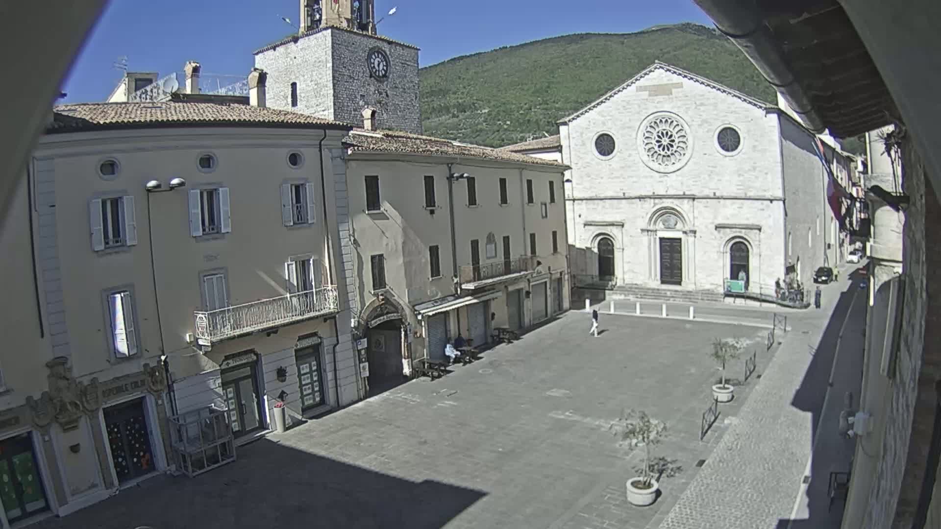 A sunny day reveals a town square with several buildings, including a church with a rose window, and a clock tower, all under a clear sky and mountains in the background.