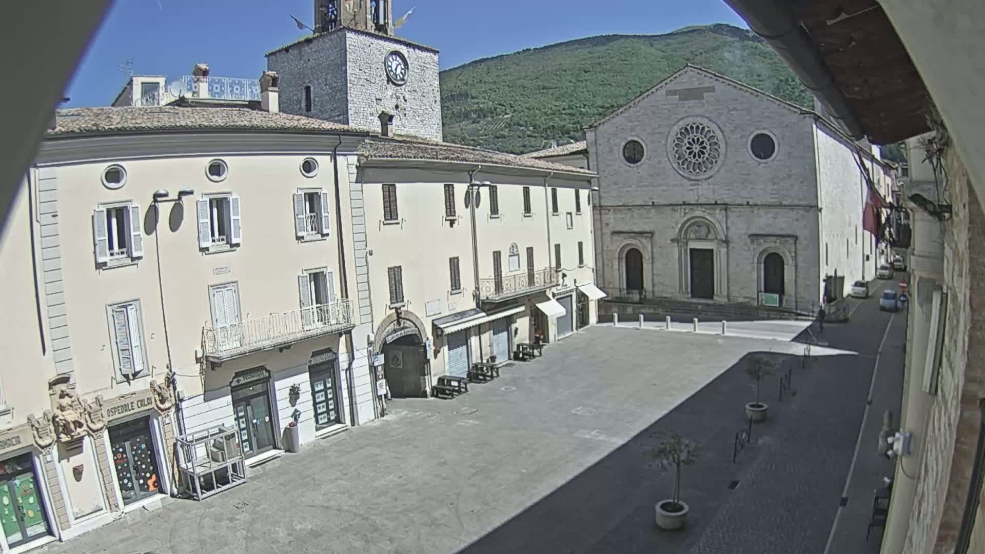 A sunny town square is surrounded by buildings, including a church with a rose window and a clock tower, under a clear sky.