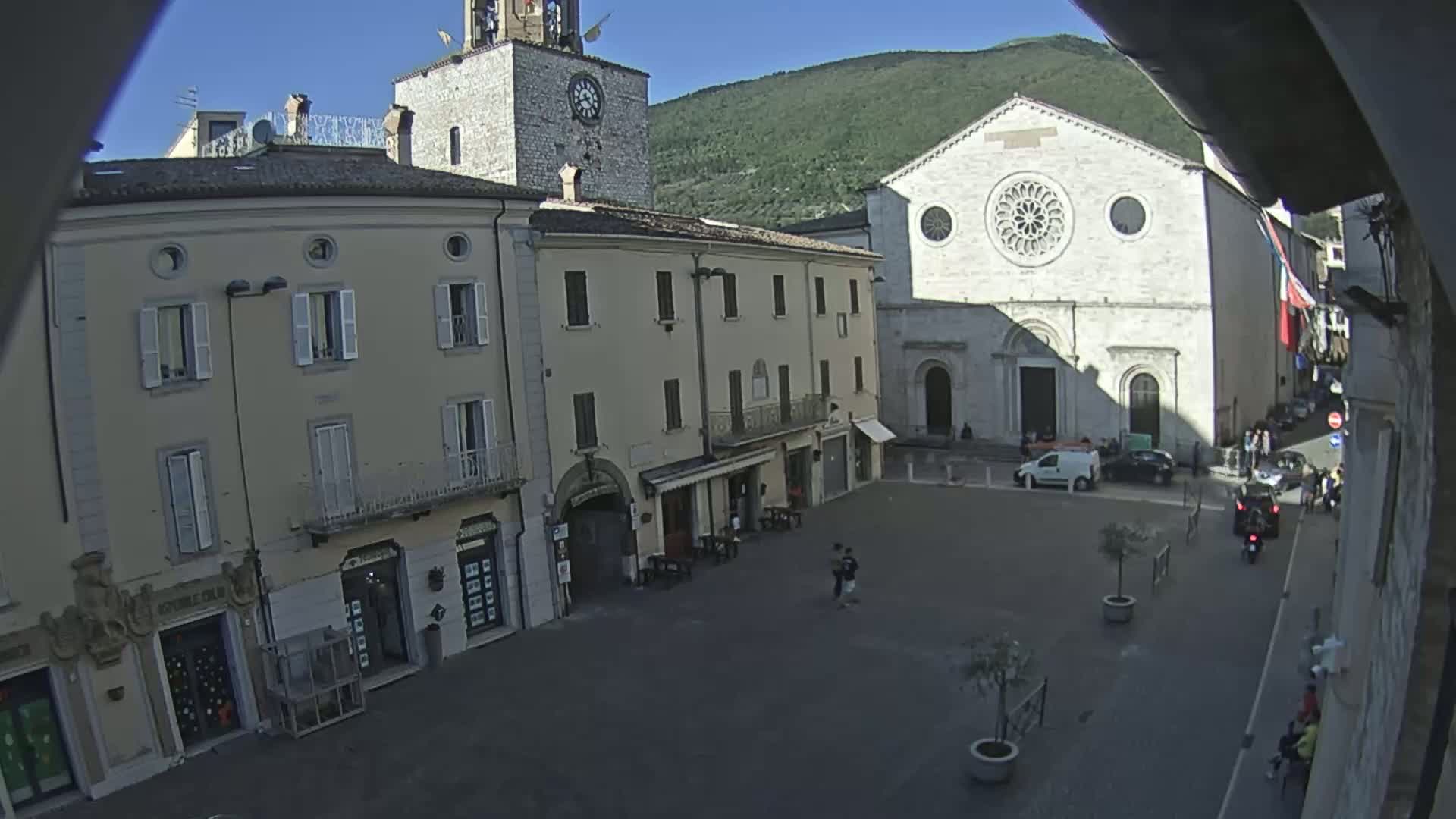 A town square is shown on a sunny day, with several multi-story buildings surrounding it, including a clock tower and a large church with a rose window.
