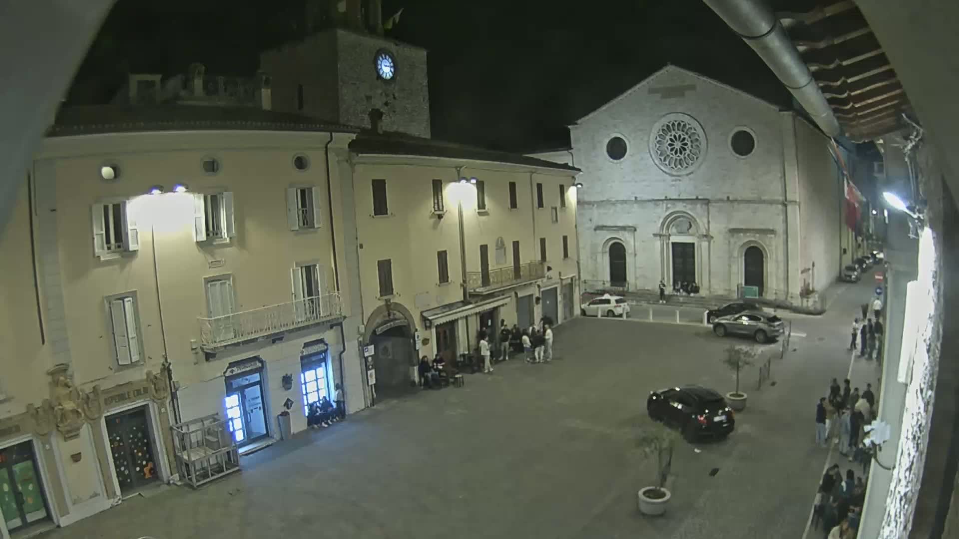 A nighttime, outdoor view of a town square with several buildings, including a church with a rose window, and people gathered in small groups.