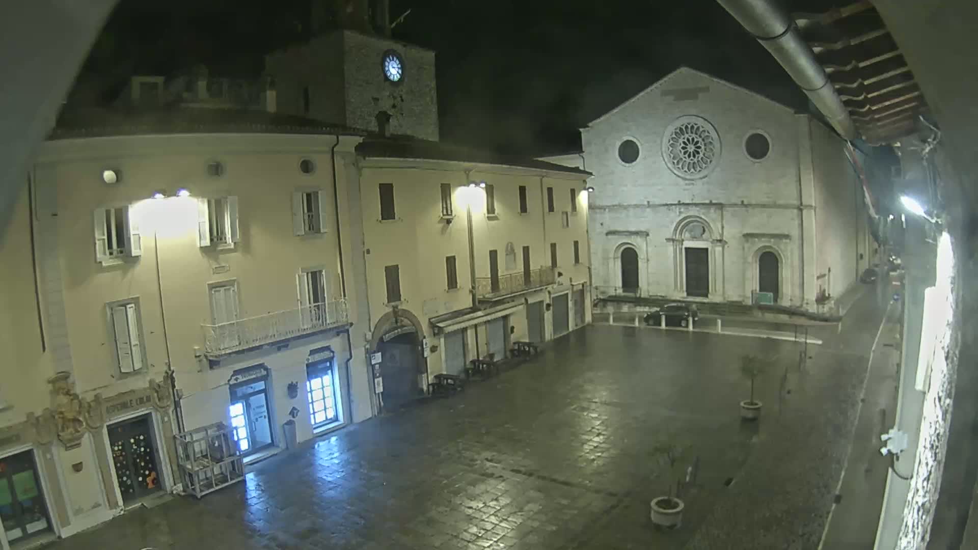 A rain-slicked town square at night is bordered by buildings, one of which features a clock tower, and another a large church with a rose window.